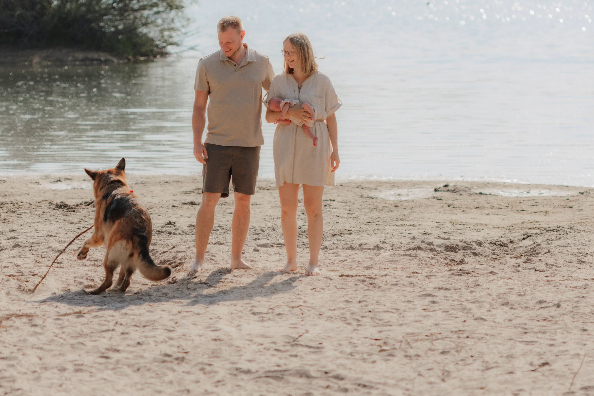 Ein Paar läuft barfuß am Strand entlang, während sie ein Baby halten, und ihr Hund spielt im Sand in der Nähe.