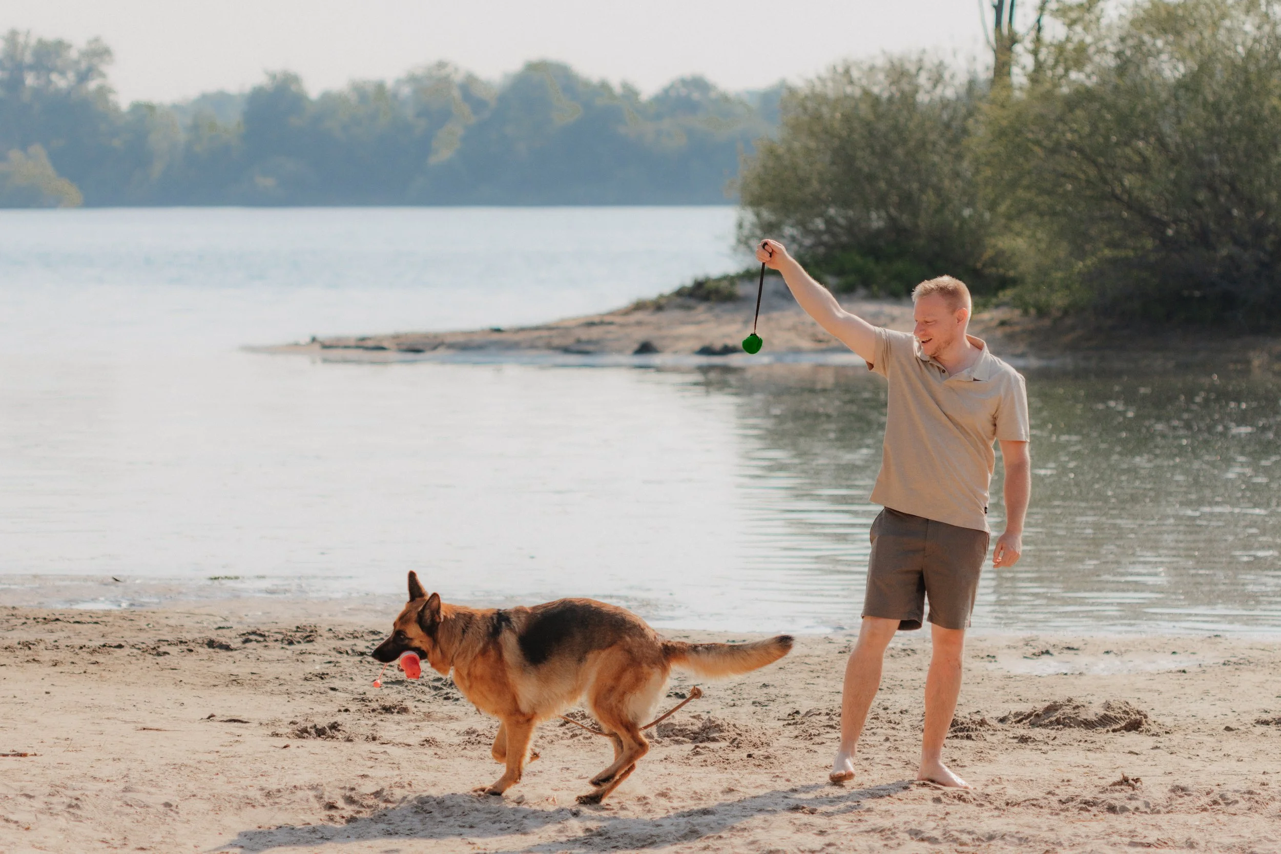 Ein Mann spielt mit seinem Hund am Strand, der Hund trägt einen roten Ball im Maul, während der Mann einen grünen Ball auffängt.
