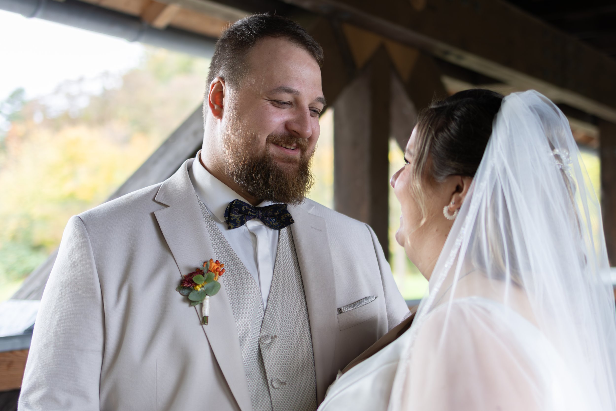 Ein braunhaariger Mann im weißen Anzug mit Fliege und Blumenboutonnière, lächelnd, schaut eine braune Frau mit Brautkleid und Schleier an, die ihn anlächelt.