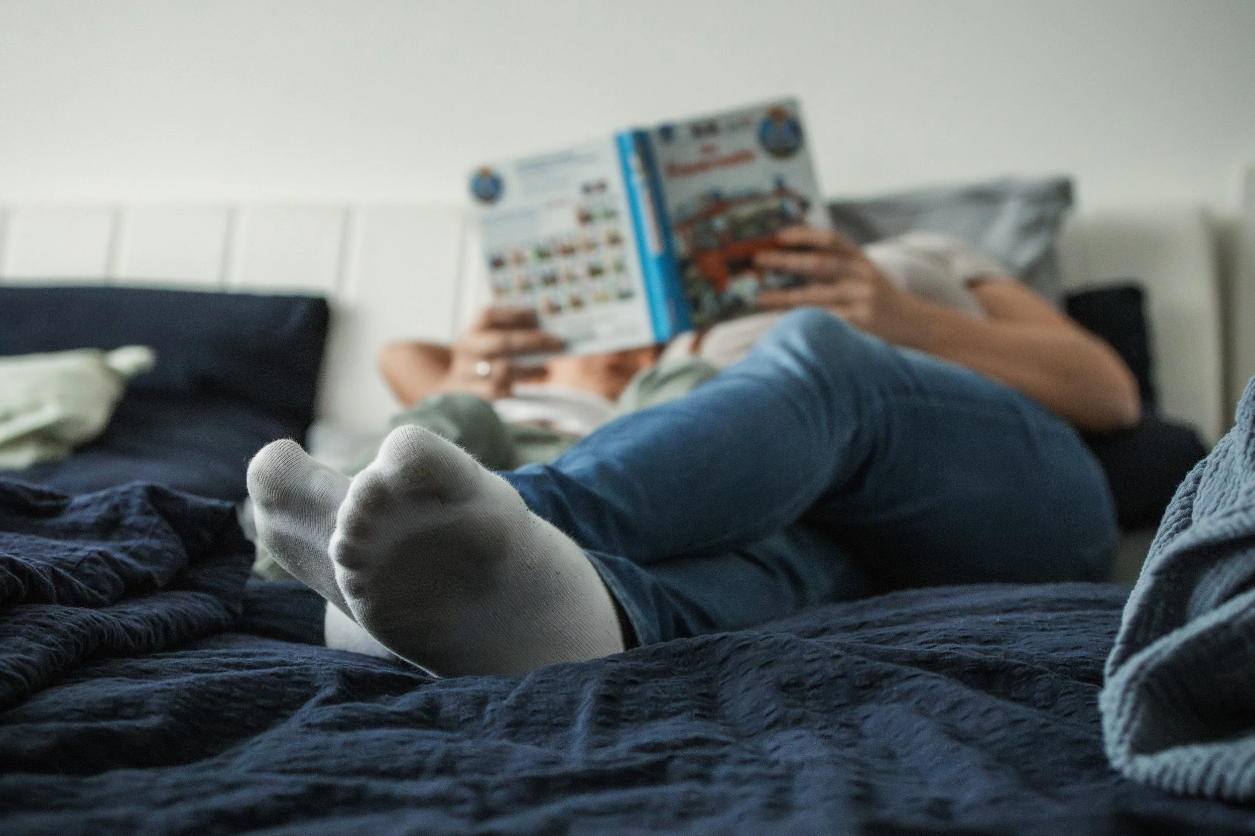 Person liegt auf einem Bett, liest ein Buch, trägt weiße Socken und Jeans, im Hintergrund ist ein Kopfteil sichtbar.
