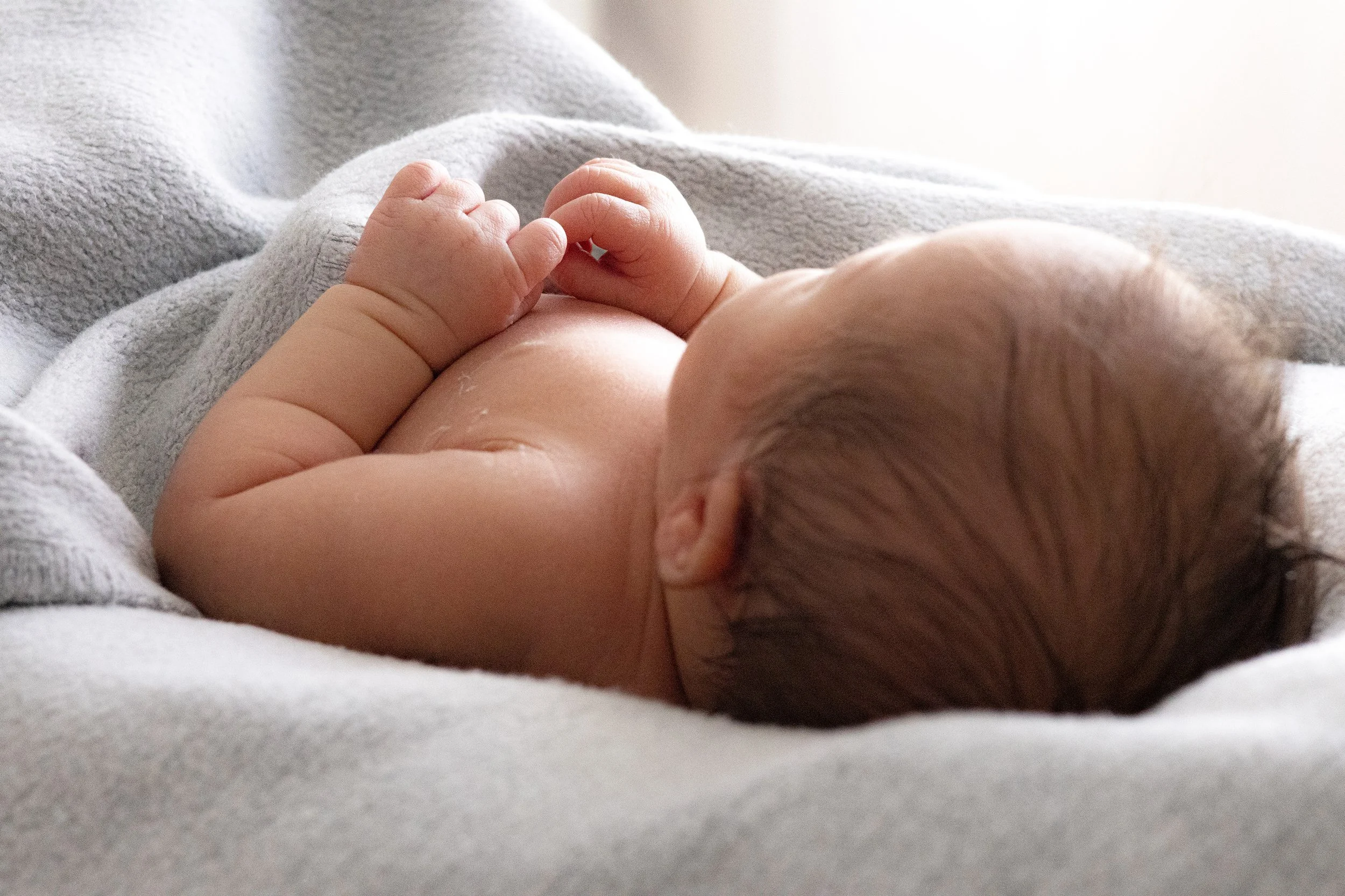 Kleines Baby liegt auf einem weichen, grauen Tuch und hält die Hände zusammen vor die Brust.