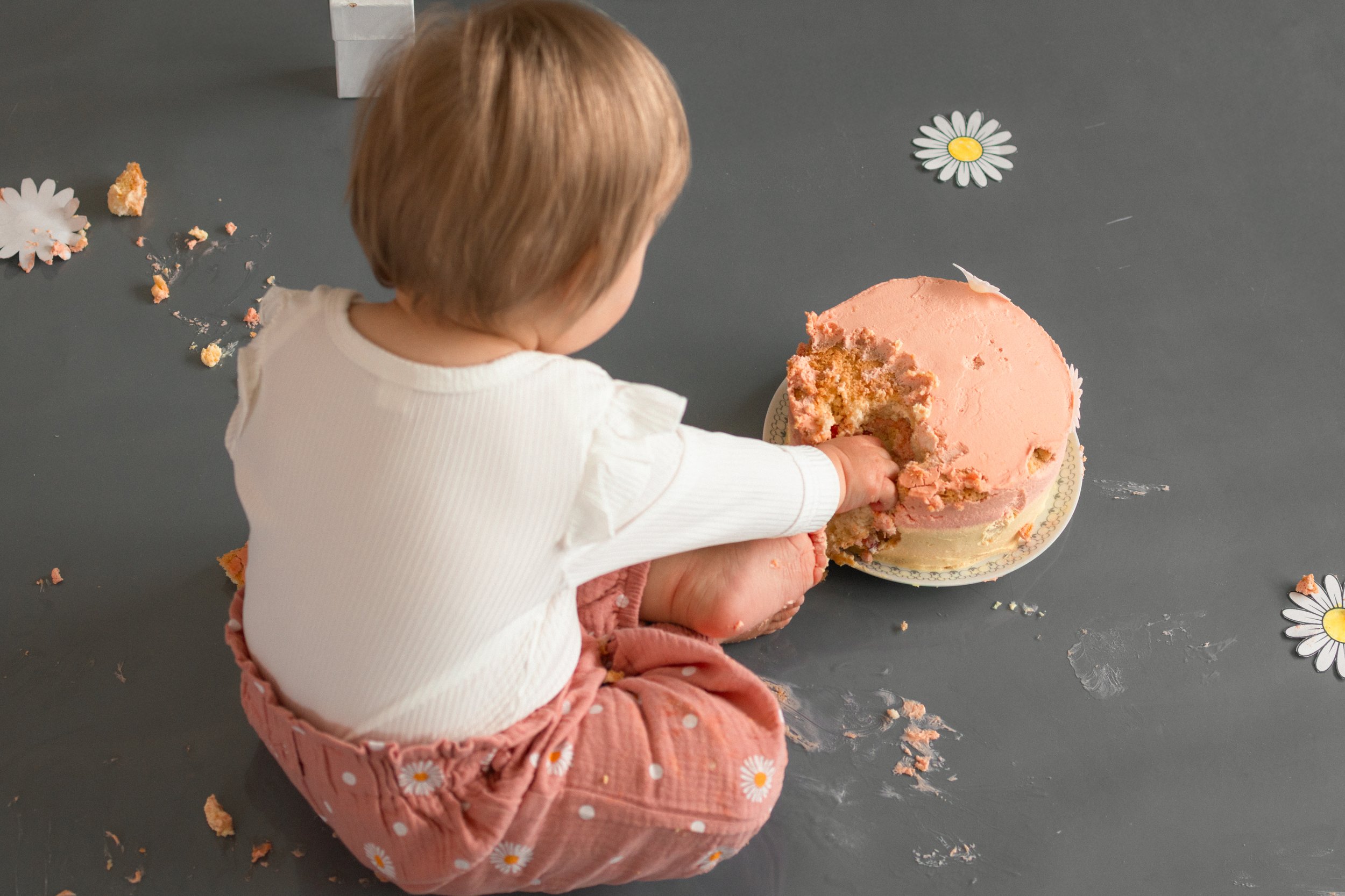 Kleines Mädchen sitzt auf dem Boden und isst eine große Torte mit rosa Glasur, umgeben von zerbröseltem Kuchen und Dekorationsblumen.