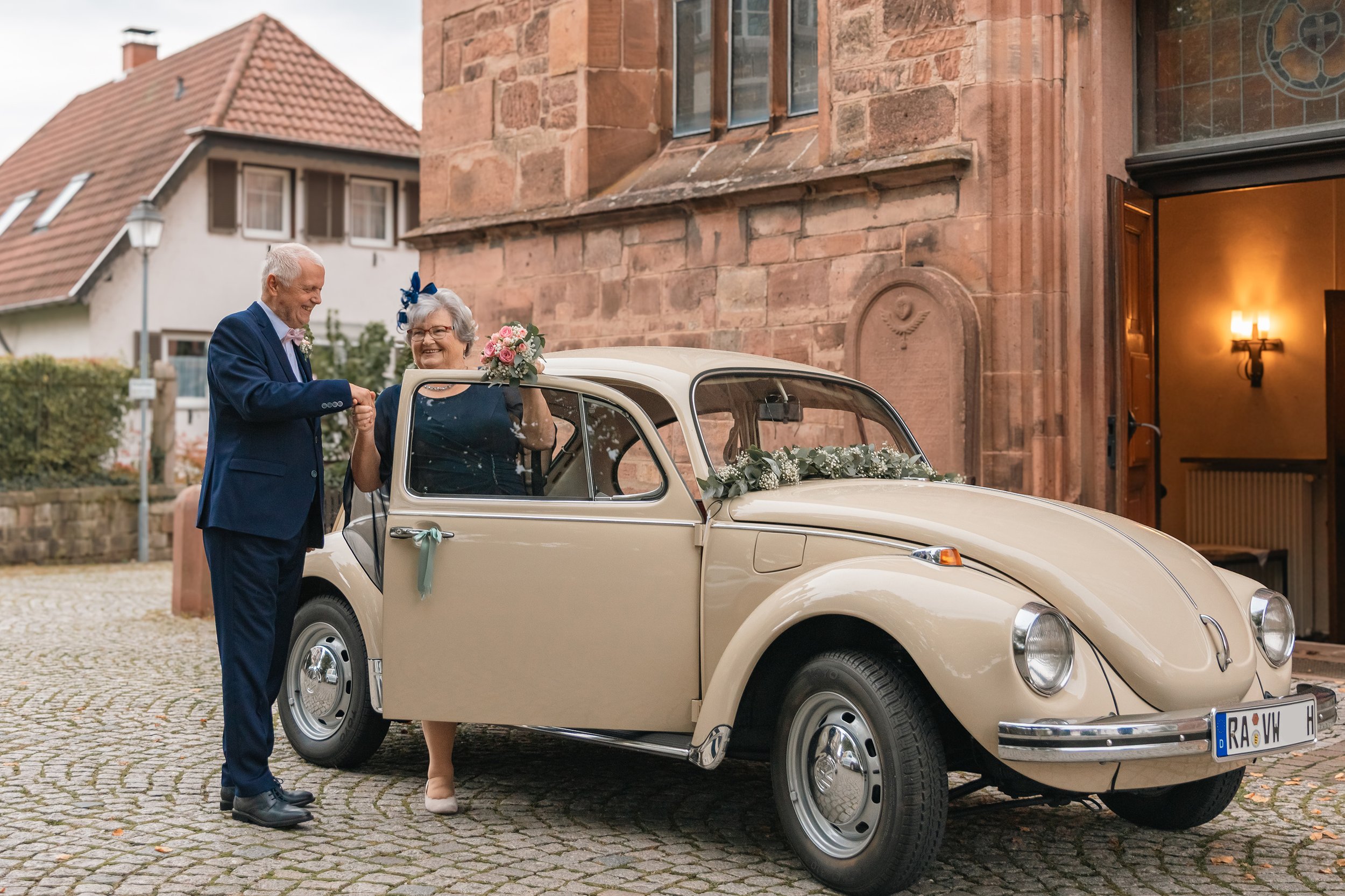 Älteres Paar bei einer Hochzeit, sie hält einen Blumenstrauß, er schüttelt ihre Hand, im Hintergrund eine alte Kirche und ein klassischer VW Käfer, der mit Blumen geschmückt ist.