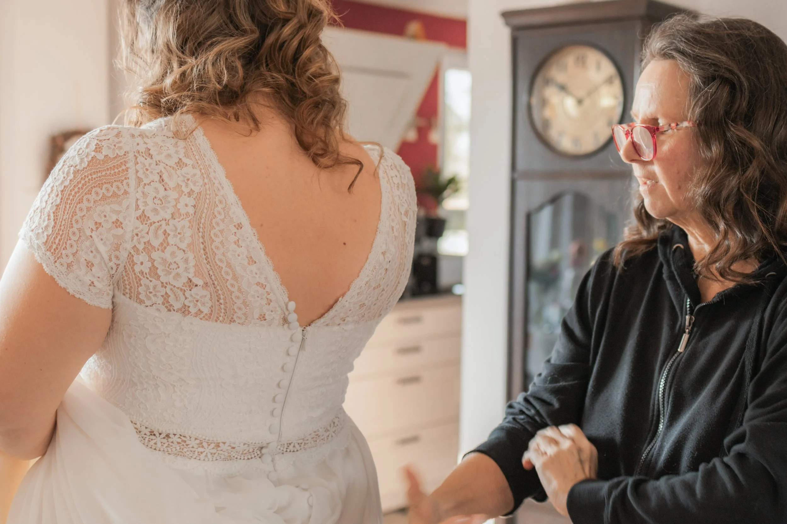 Eine Braut in einem weißen Kleid mit Spitze wird von einer älteren Frau mit Brille im schwarzen Jackett in einem freundlichen Moment betreut.
