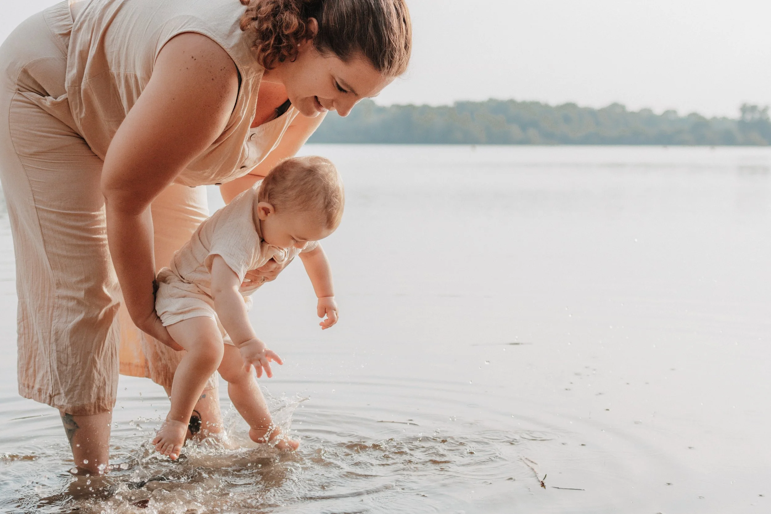 Eine Frau hält ein Kleinkind an den Beinen am Wasserstand, während es ins Wasser greift, bei Sonnenuntergang oder -dämmerung am Ufer eines Sees.