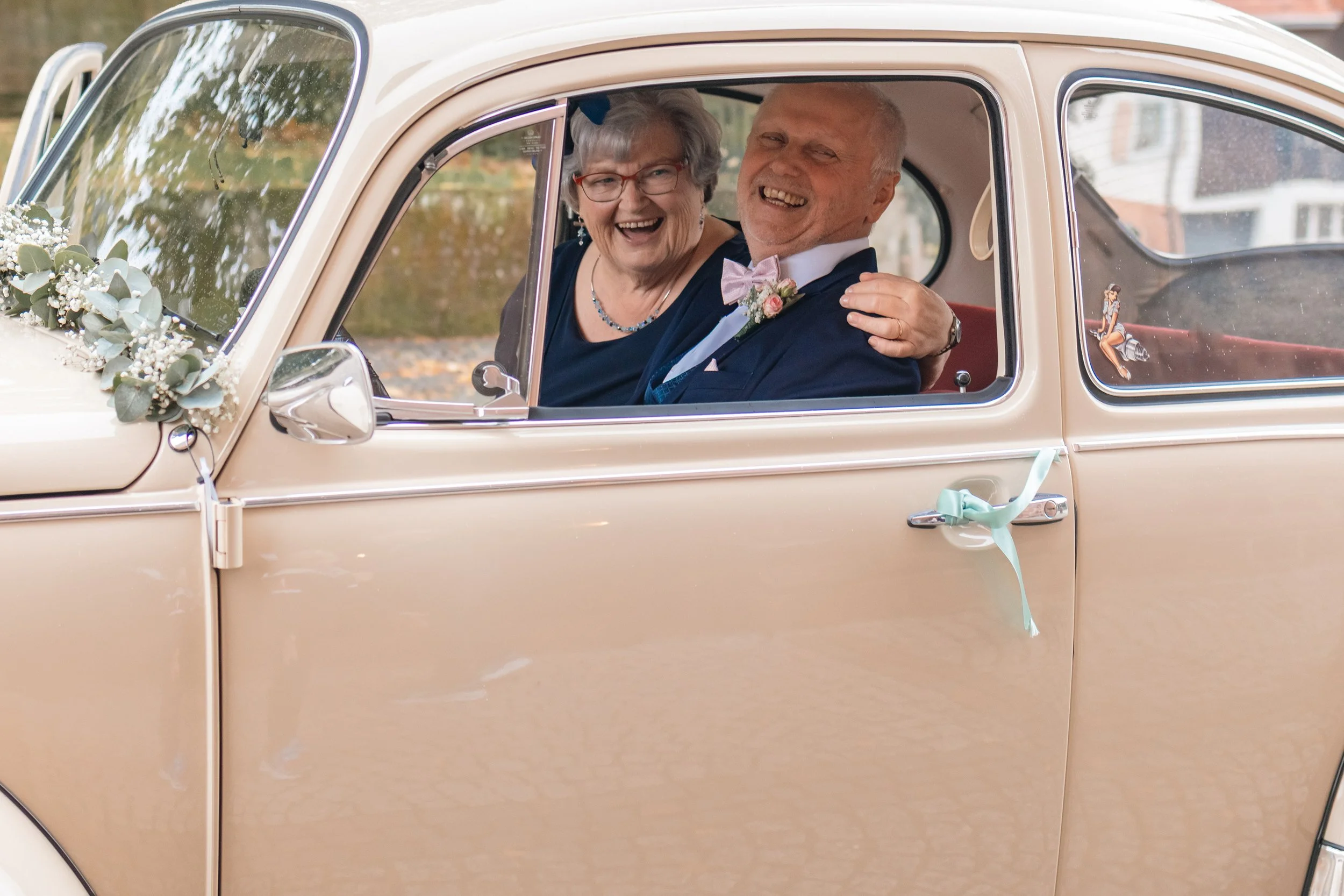 Ein älteres Paar in einem vintage beige Auto, das wahrscheinlich für eine Hochzeit dekoriert ist, lächelt lachend. Der Mann trägt einen Anzug mit Fliege und eine Blume am Revers, die Frau trägt eine dunkelblaue Kleidung und Brille.