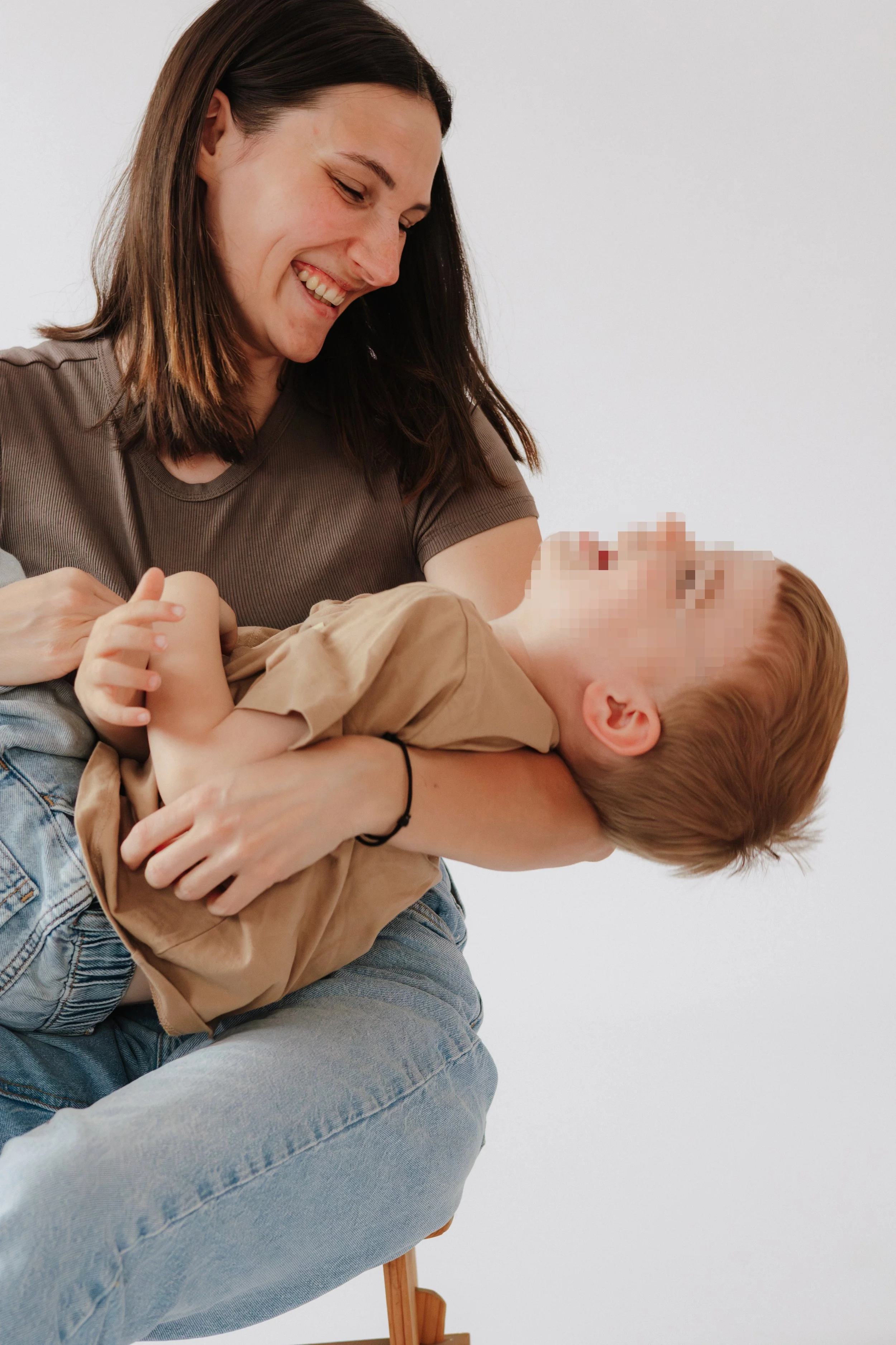 Eine junge Frau hält ein lachendes Kind in ihren Armen, das auf einem Stuhl sitzt. Das Kind trägt einen beigen Pullover. Die Frau trägt ein braunes T-Shirt und blaue Jeans. Sie lachen beide und die Szene spielt vor einem weißen Hintergrund.