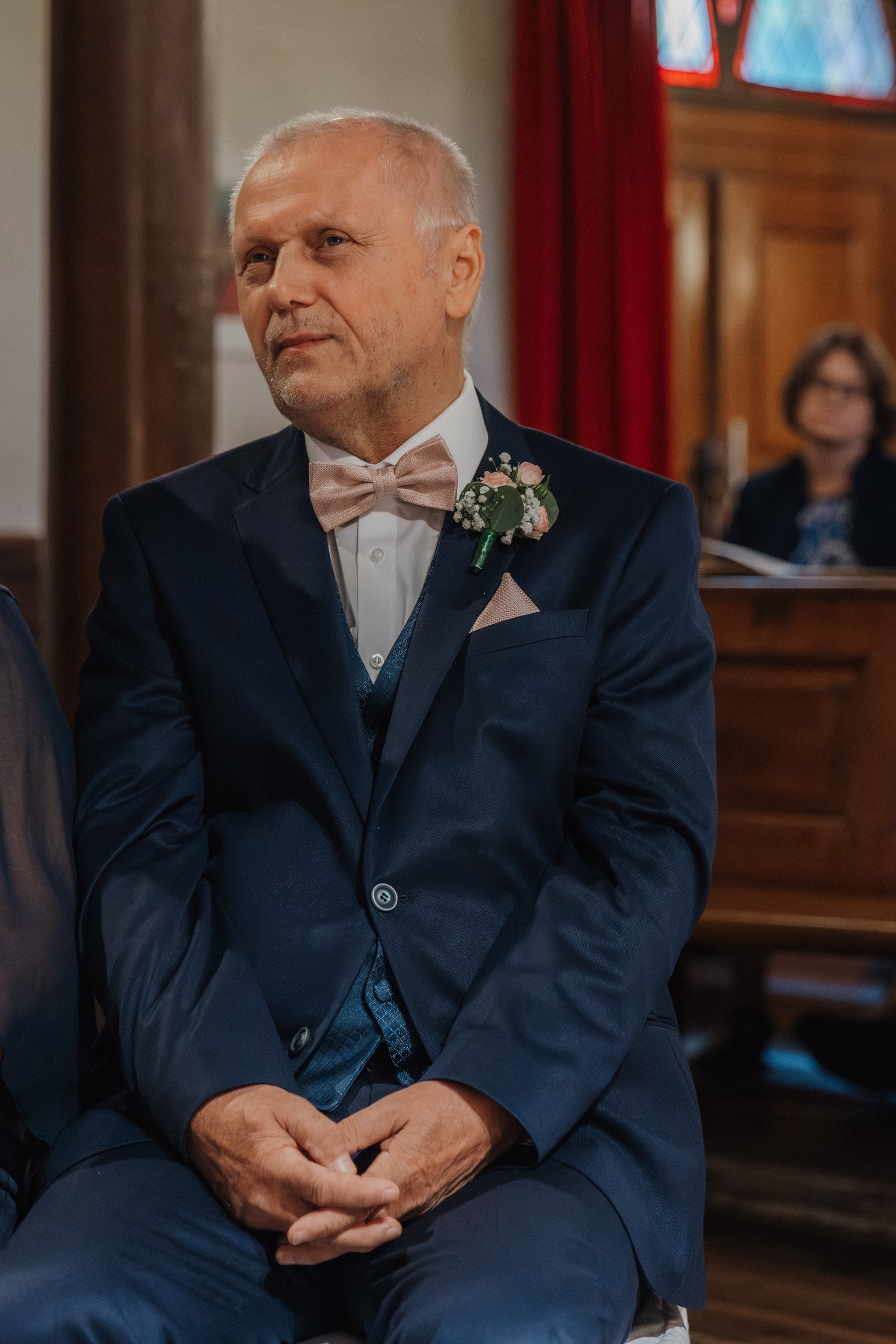 Ein älterer Mann in einem dunklen Anzug mit weißen Hemd, rosa Fliege und Blumenanstecker sitzt bei einer Hochzeitsfeier in einer Kirche.