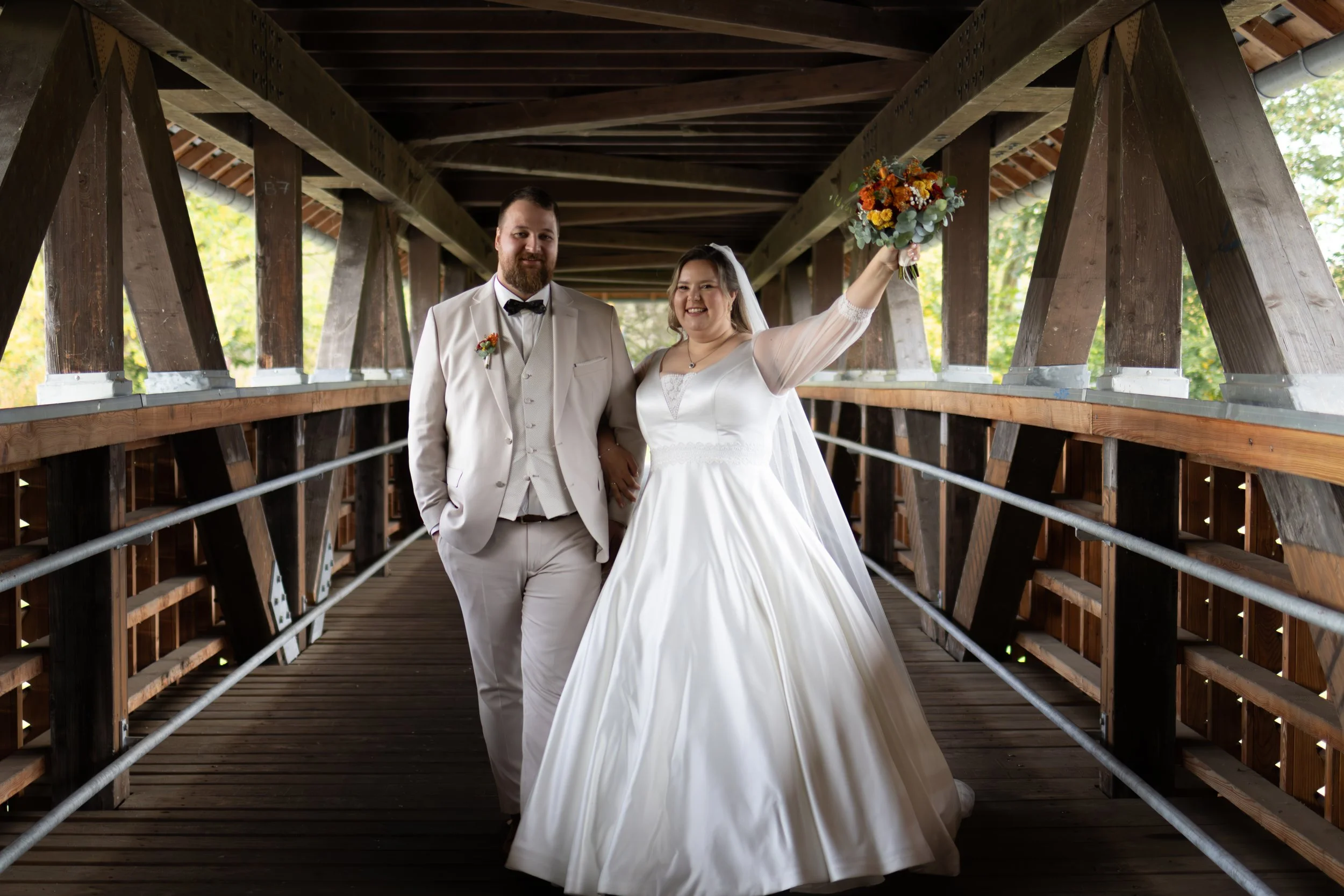 Ein Brautpaar in Hochzeitskleidung auf einer Holzbrücke, die Braut hält einen Blumenstrauß hoch, beide lächeln, im Hintergrund sind Bäume und natürliches Licht.