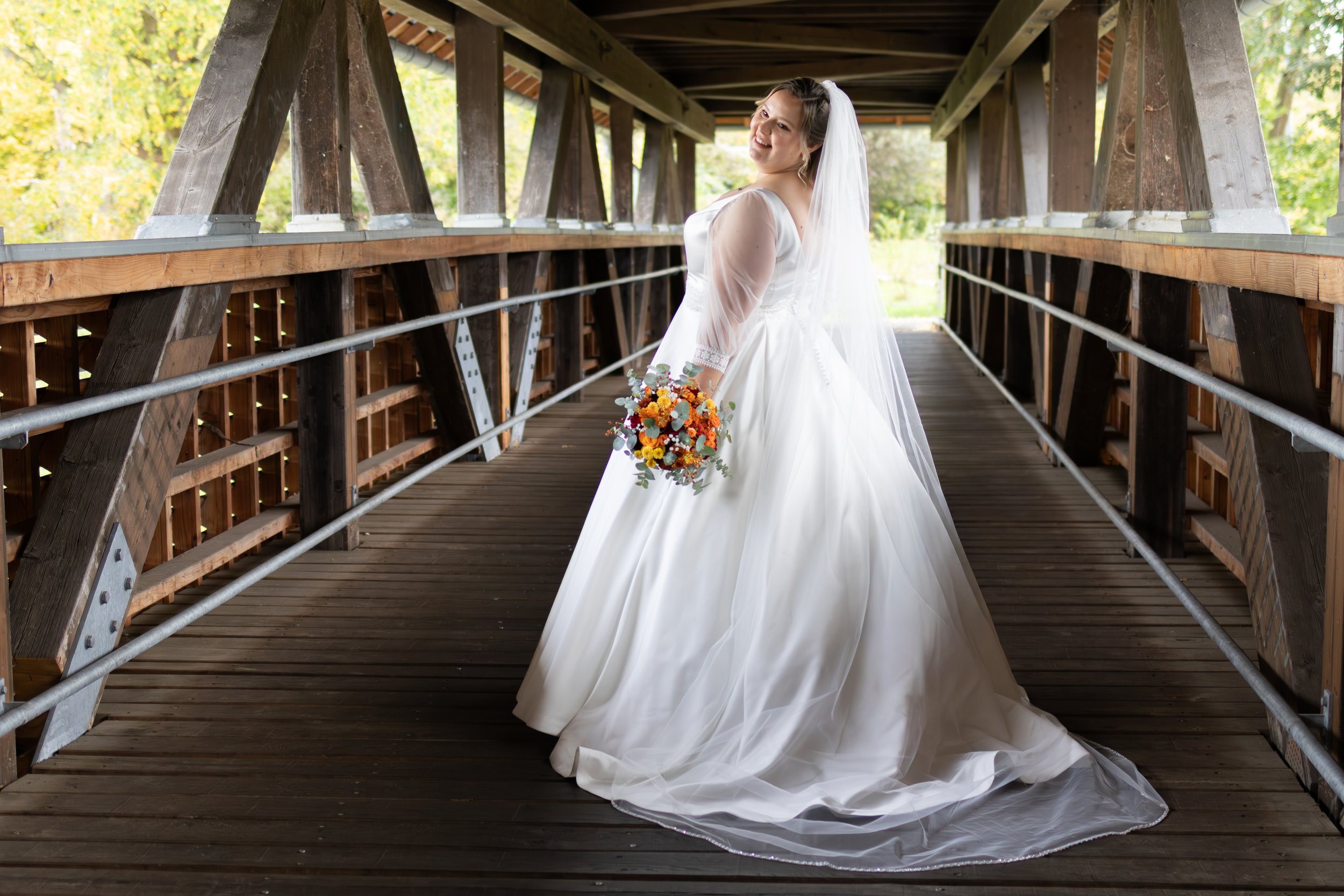 Braut in weißem Hochzeitskleid mit Blumenstrauß auf einer Holzbrücke, umgeben von Natur.