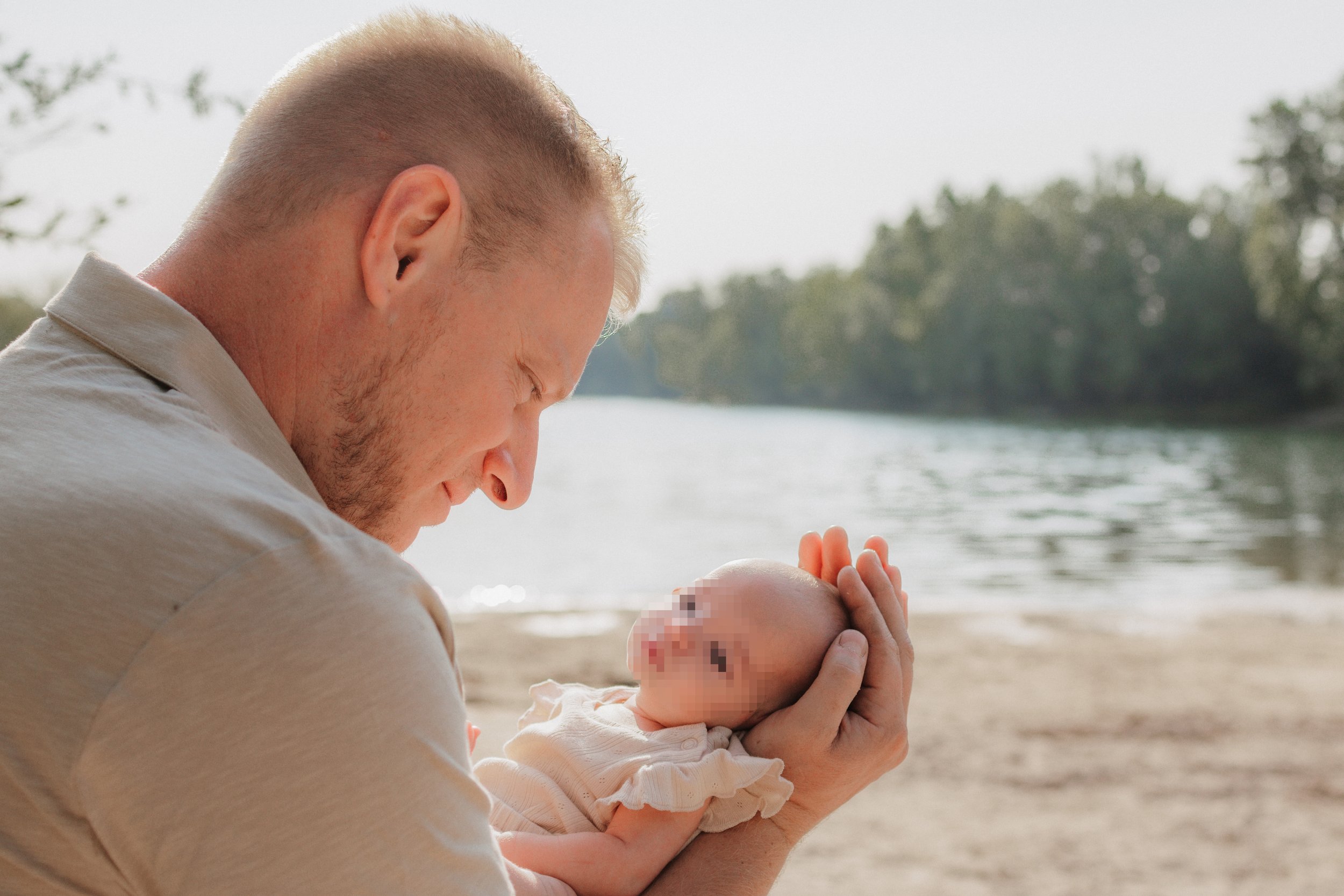 Vater hält Baby am Flussufer, während sie sich liebevoll anschauen, im Hintergrund Wasser und Bäume, sonniger Tag.