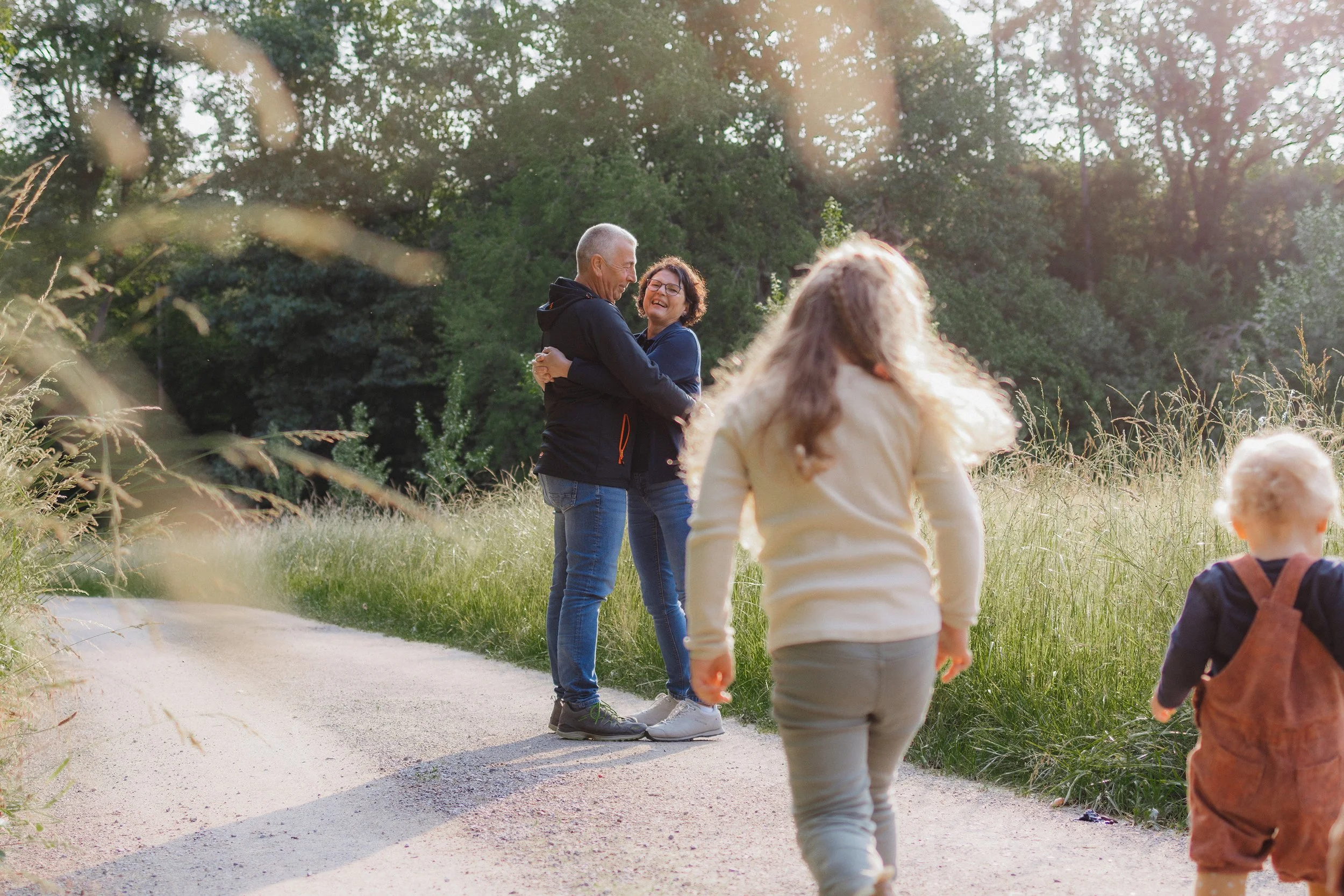 Eine Familie mit Kindern spaziert in einem Park, im Hintergrund ein Erwachsen Paar, das sich umarmt und lacht, bei Sonnenuntergang.