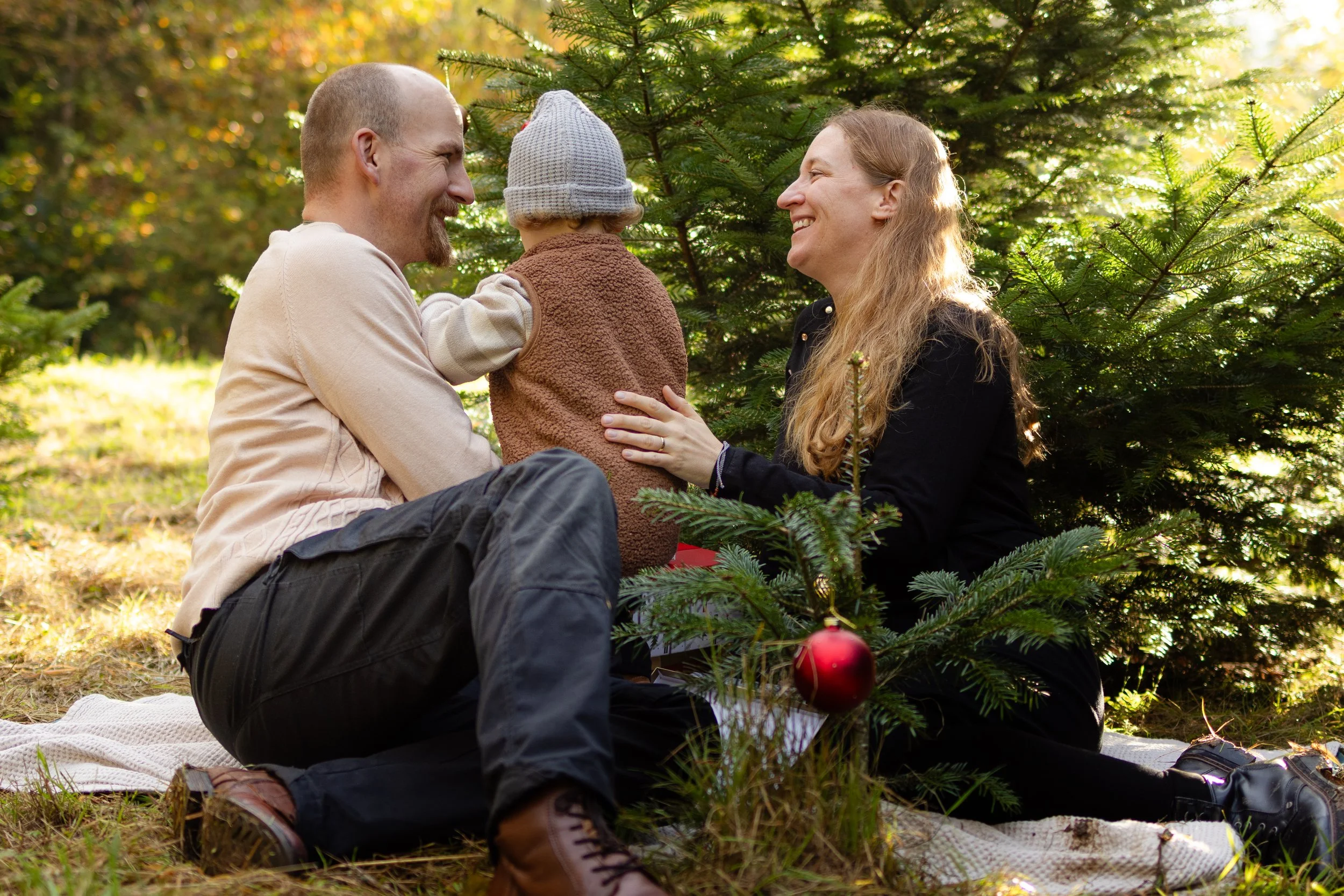 Eine glückliche Familie sitzt im Wald neben einem Weihnachtsbaum, der mit einer roten Weihnachtskugel dekoriert ist, während die Mutter und der Vater mit ihrem Kind lachen.