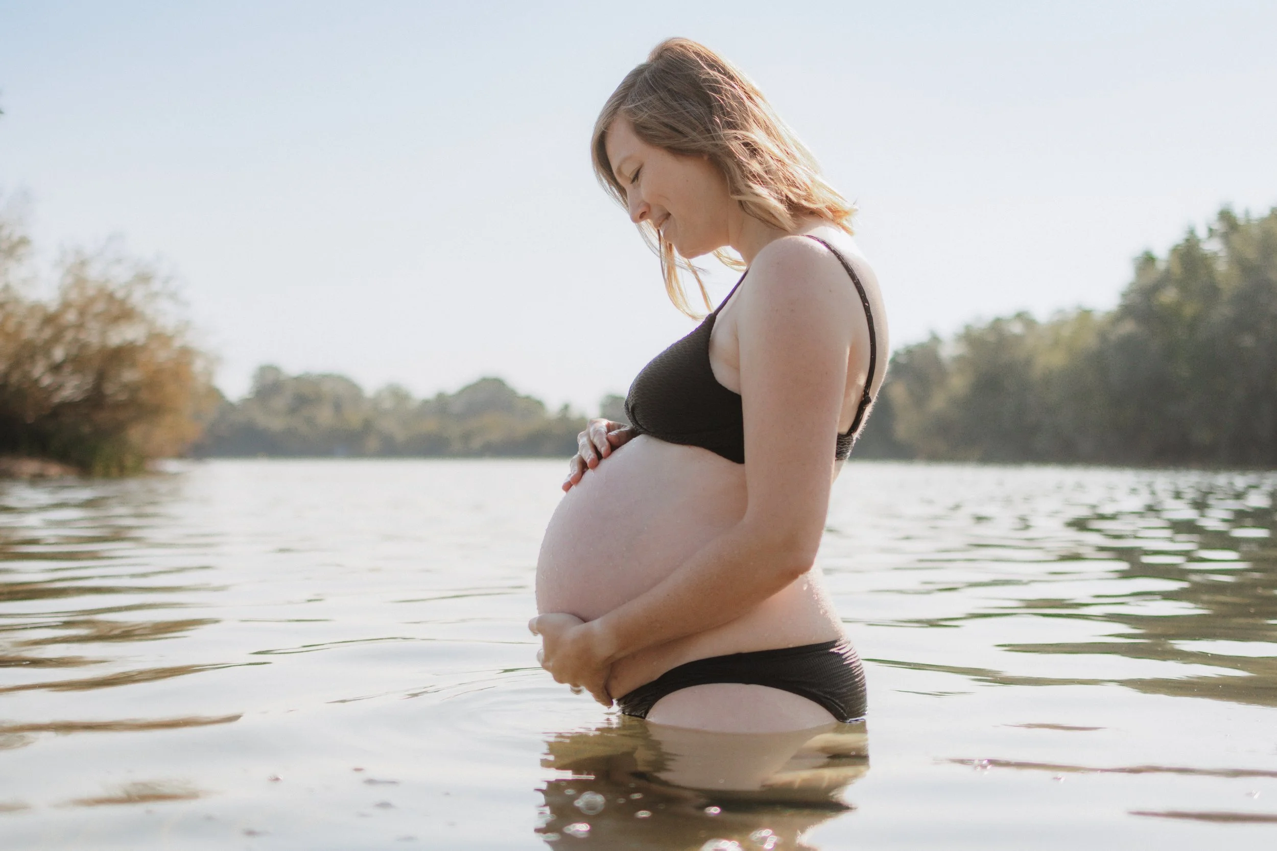 Schwangere Frau im Wasser, die ihre Schwangerschaft sichtbar macht, bei Sonnenlicht am Fluss mit Bäumen im Hintergrund.