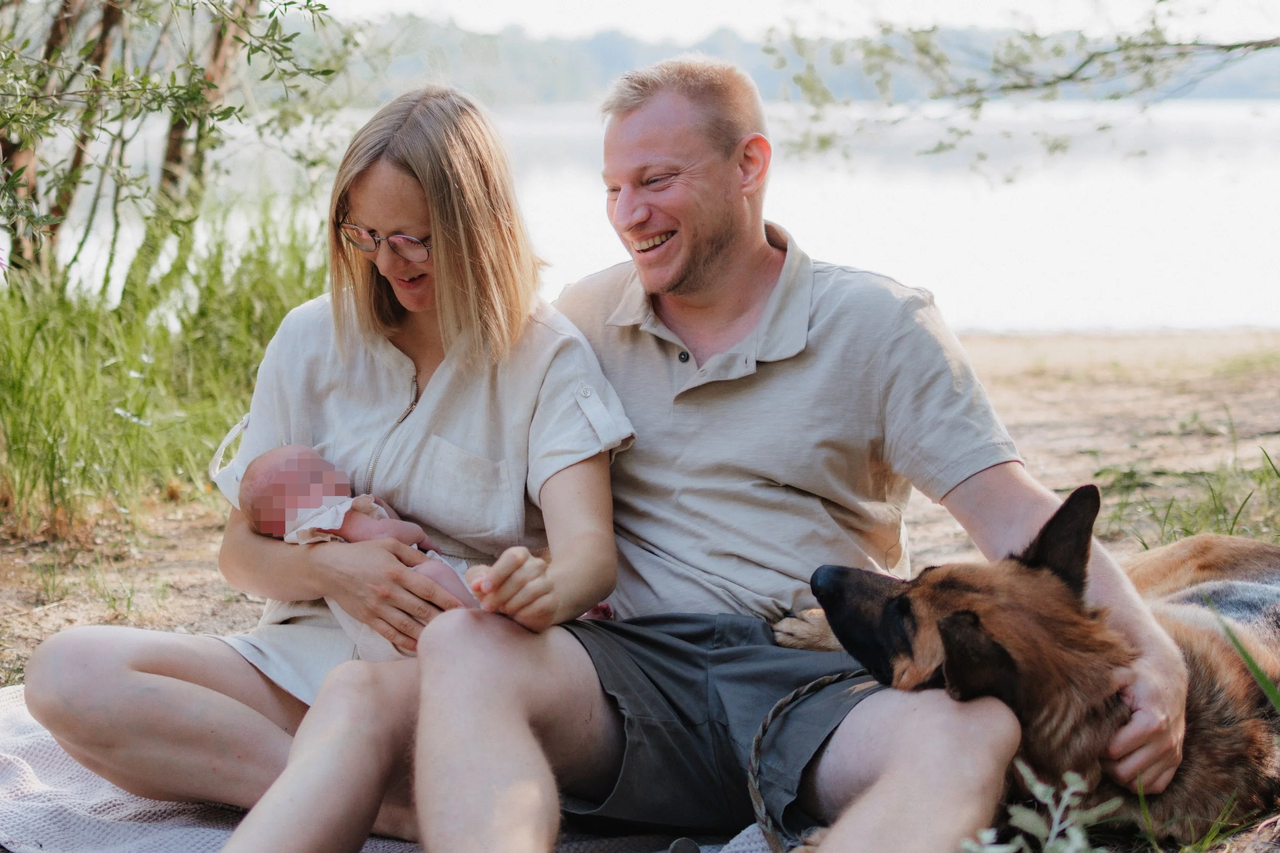 Ein glückliches Paar sitzt am Strand unter Bäumen, die Mutter hält ein neugeborenes Baby, während ein Hund neben ihnen liegt.