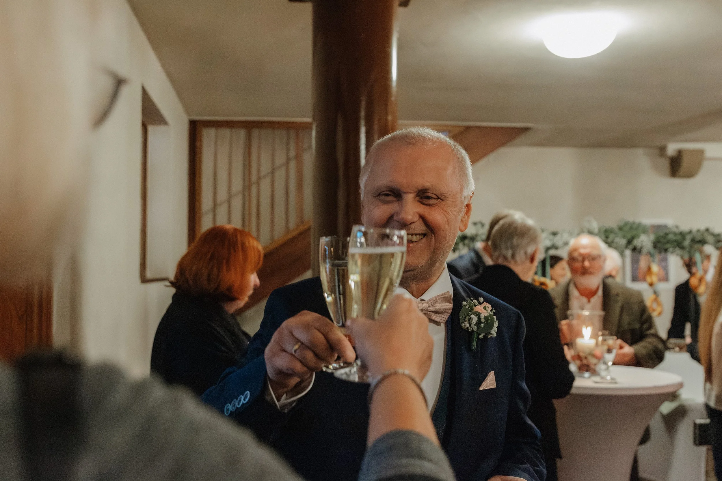 Ein älterer Mann in formeller Kleidung, der bei einer Feier mit einem Glas Sekt anstößt, lächelnd.