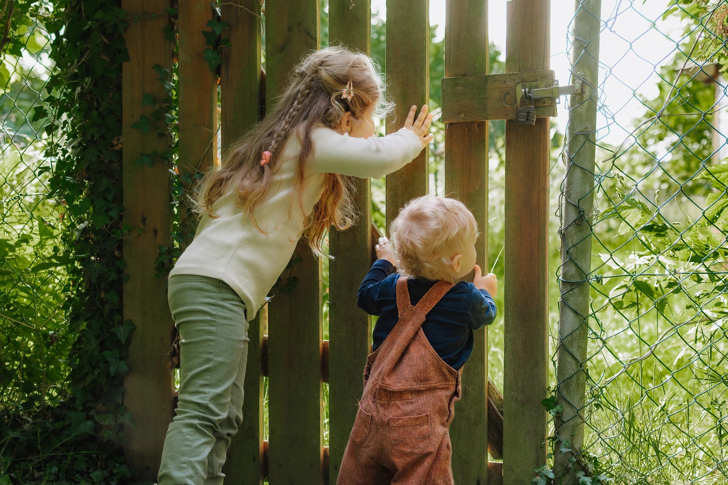 Zwei Kinder, ein Mädchen mit langem Haar und ein jüngeres Kind, schauen durch ein Gartengitter.