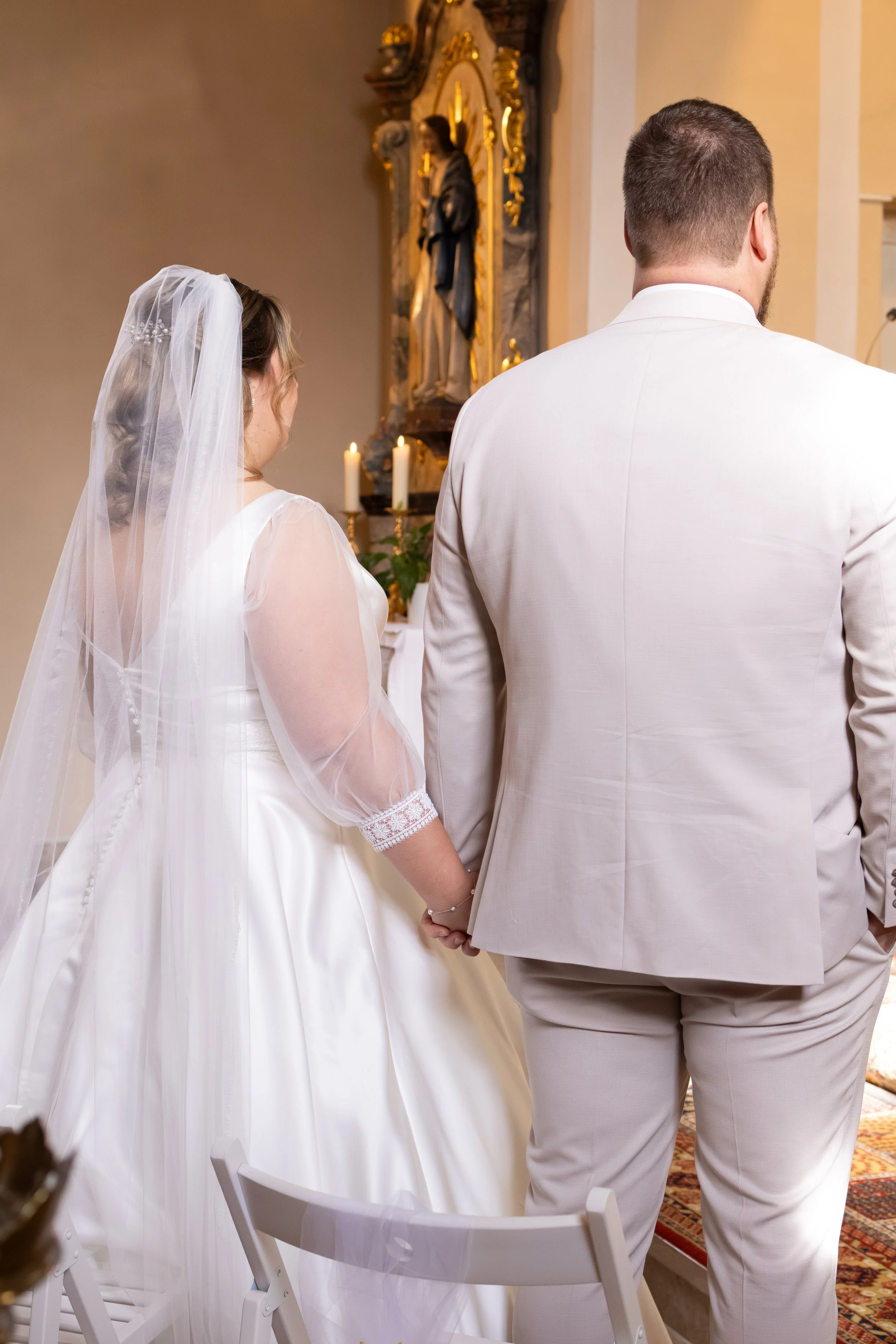 Braut und Bräutigam bei einer Hochzeit in einer Kirche, vom Rücken gesehen, die Hand halten, mit religiösen Figuren und Kerzen im Hintergrund.