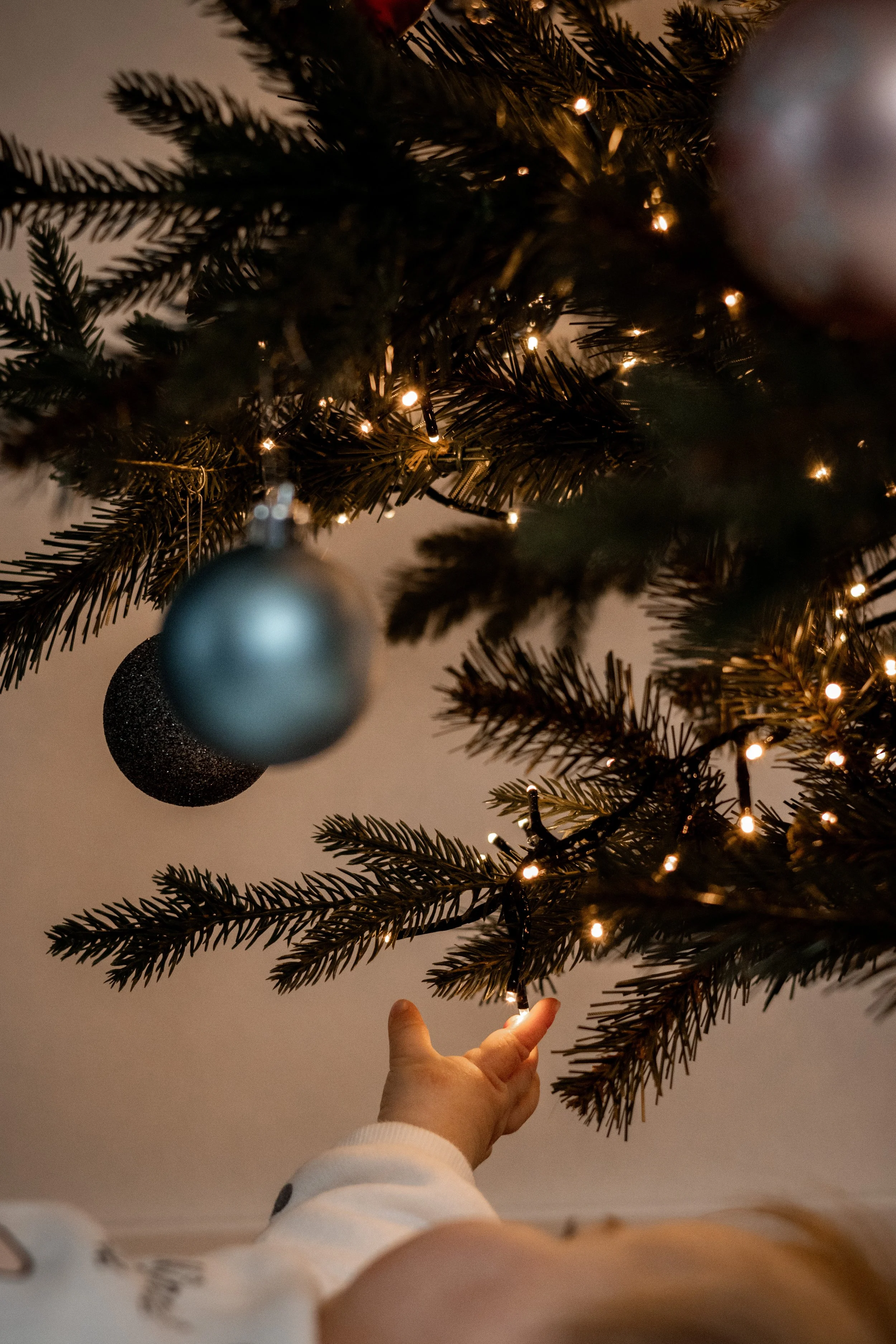 Ein Weihnachtsbaum mit Lichterketten und Kugeln, darunter eine Hand, die an einem Ast berührt.