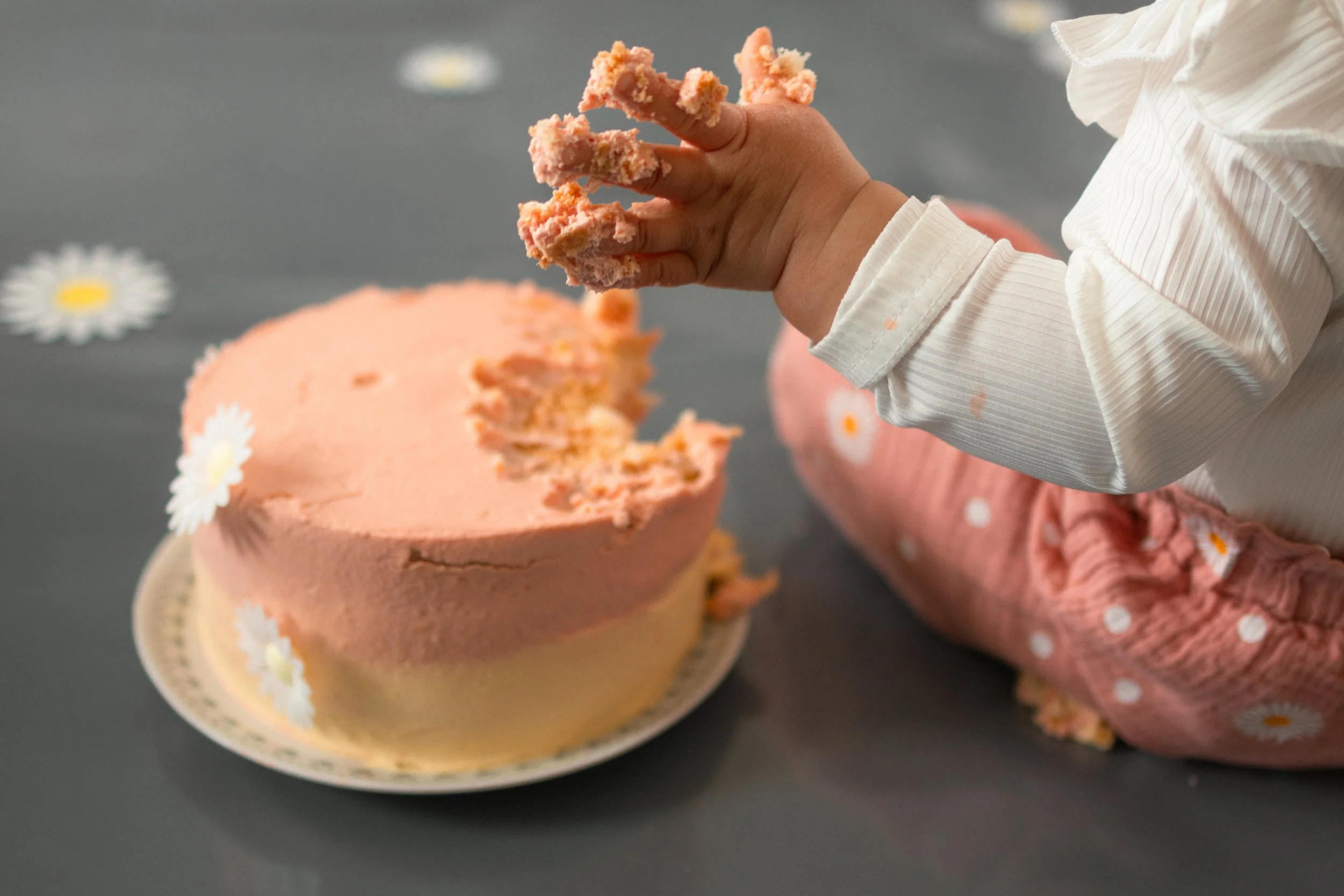 Kleinkind kratzt an einer rosa Torte mit daisy-Dekor, die auf einem Teller steht, während es auf dem Boden liegt.