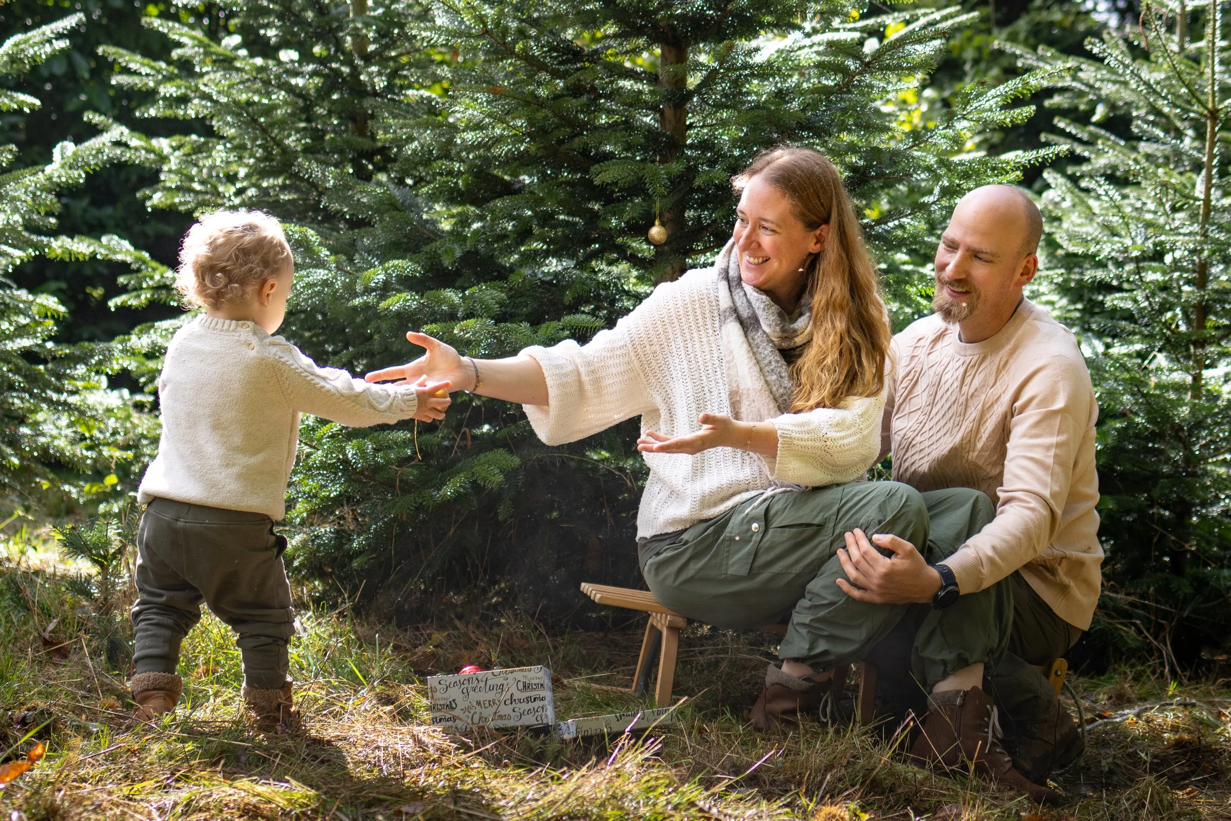 Familie sitzt im Wald vor Weihnachtsbäumen, Kind erhält ein Geschenk, Mama reicht es, Papa unterstützt, alles ist fröhlich und festlich.