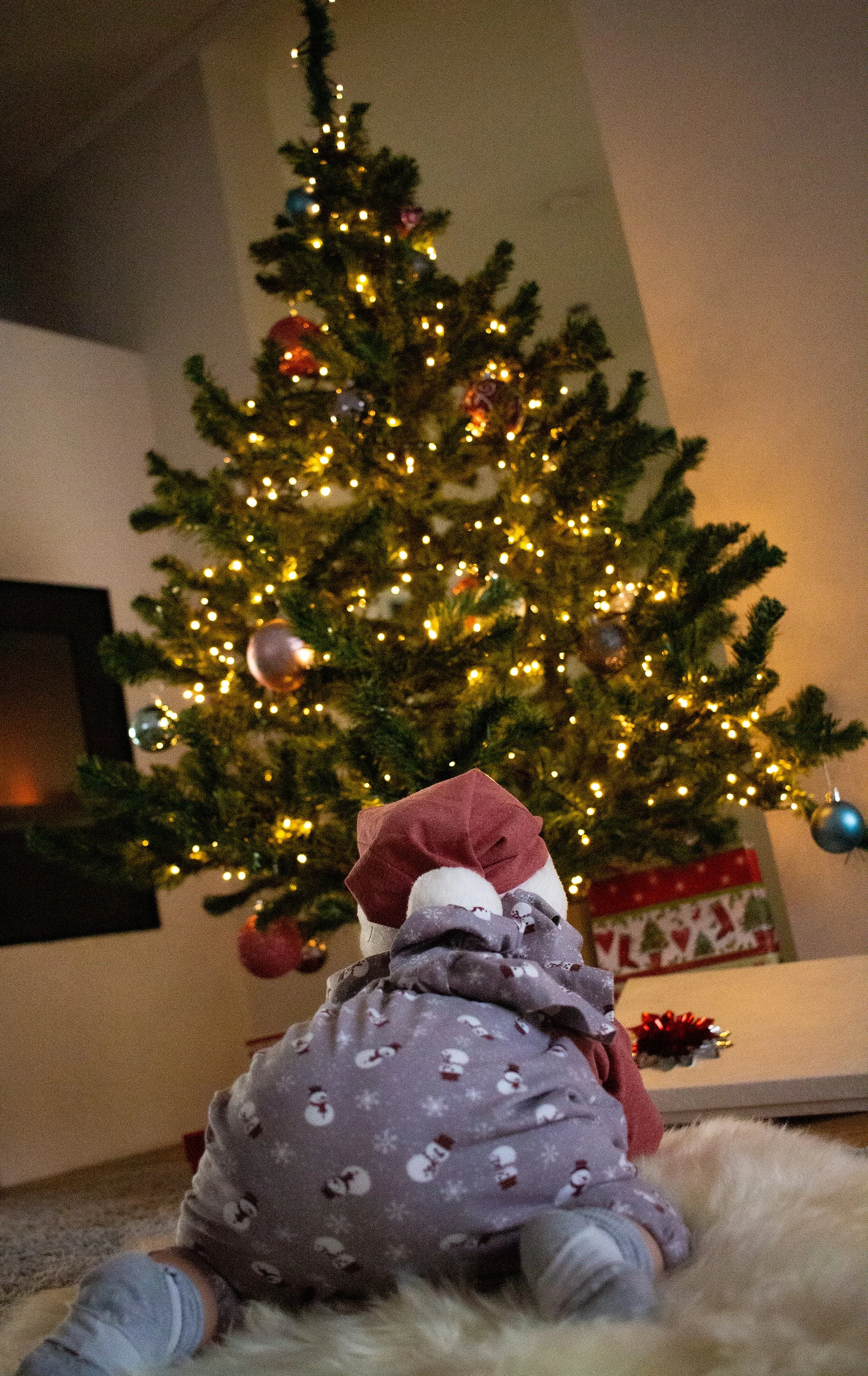 Kleines Kind in Weihnachtskleidung sitzt vor einem geschmückten Weihnachtsbaum.