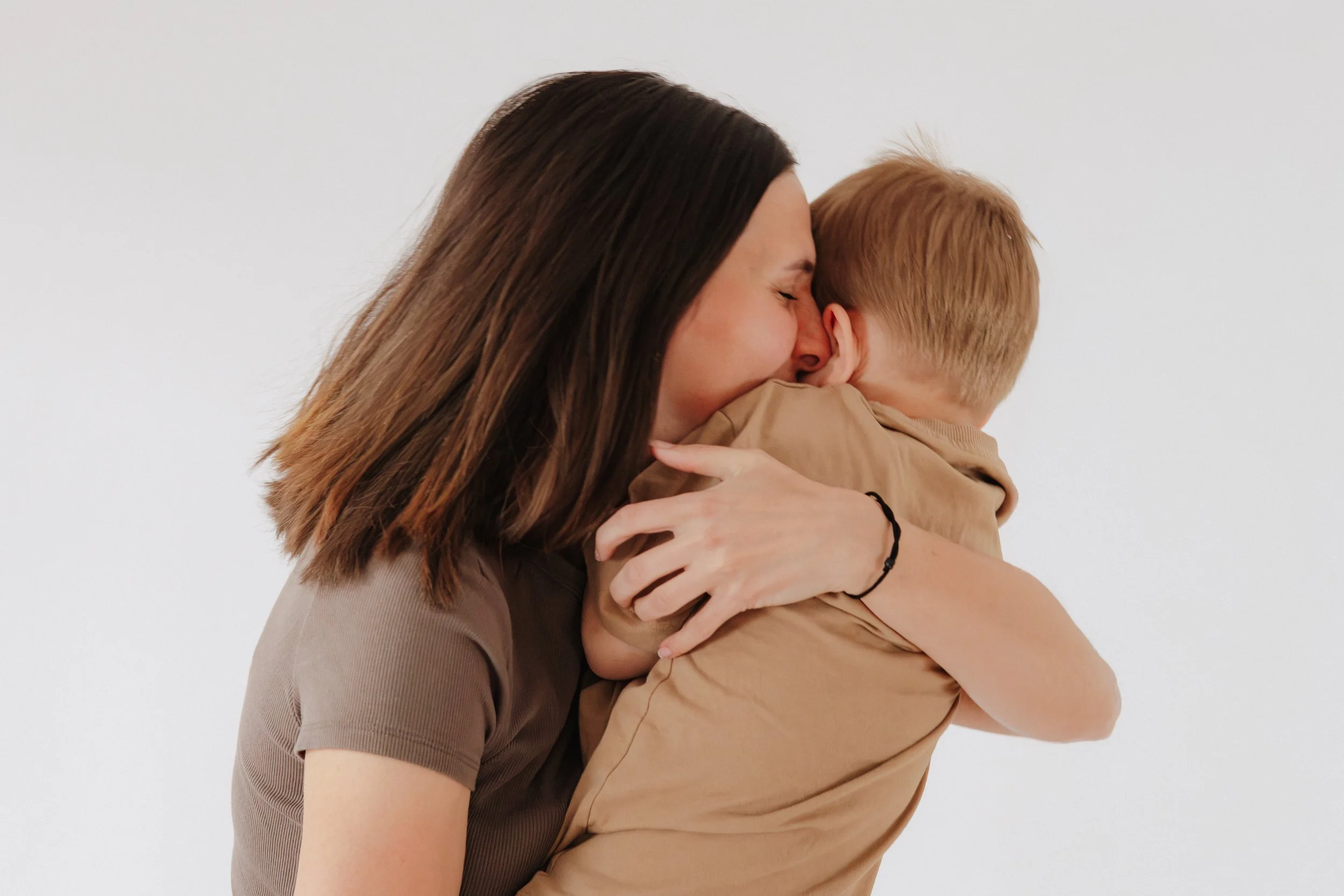 Eine Frau umarmt einen kleinen Jungen, beide haben die Augen geschlossen und lächeln. Die Szene ist liebevoll und herzlich, vor einem weißen Hintergrund.