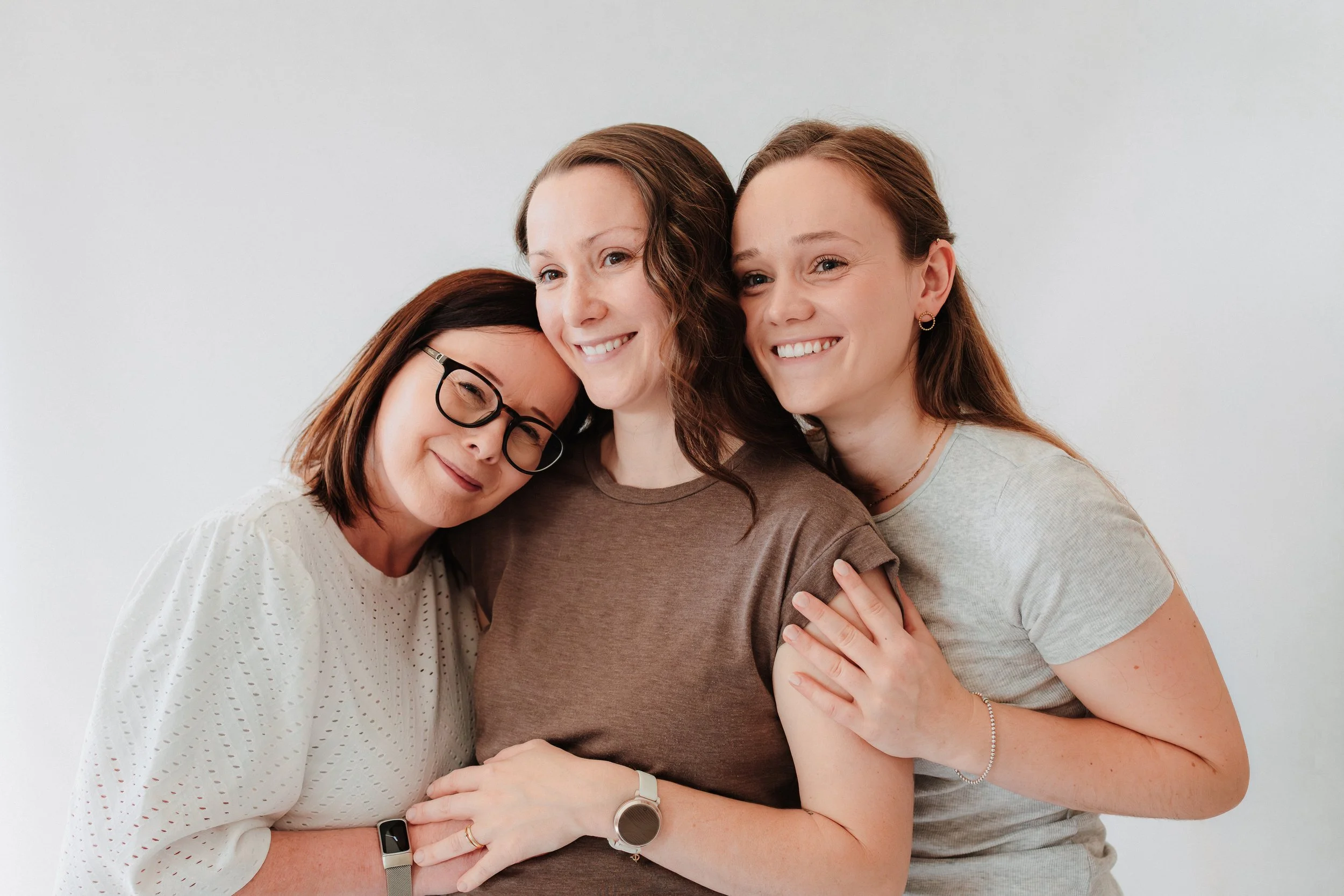 Drei Frauen umarmen sich und lachen vor einem weißen Hintergrund.