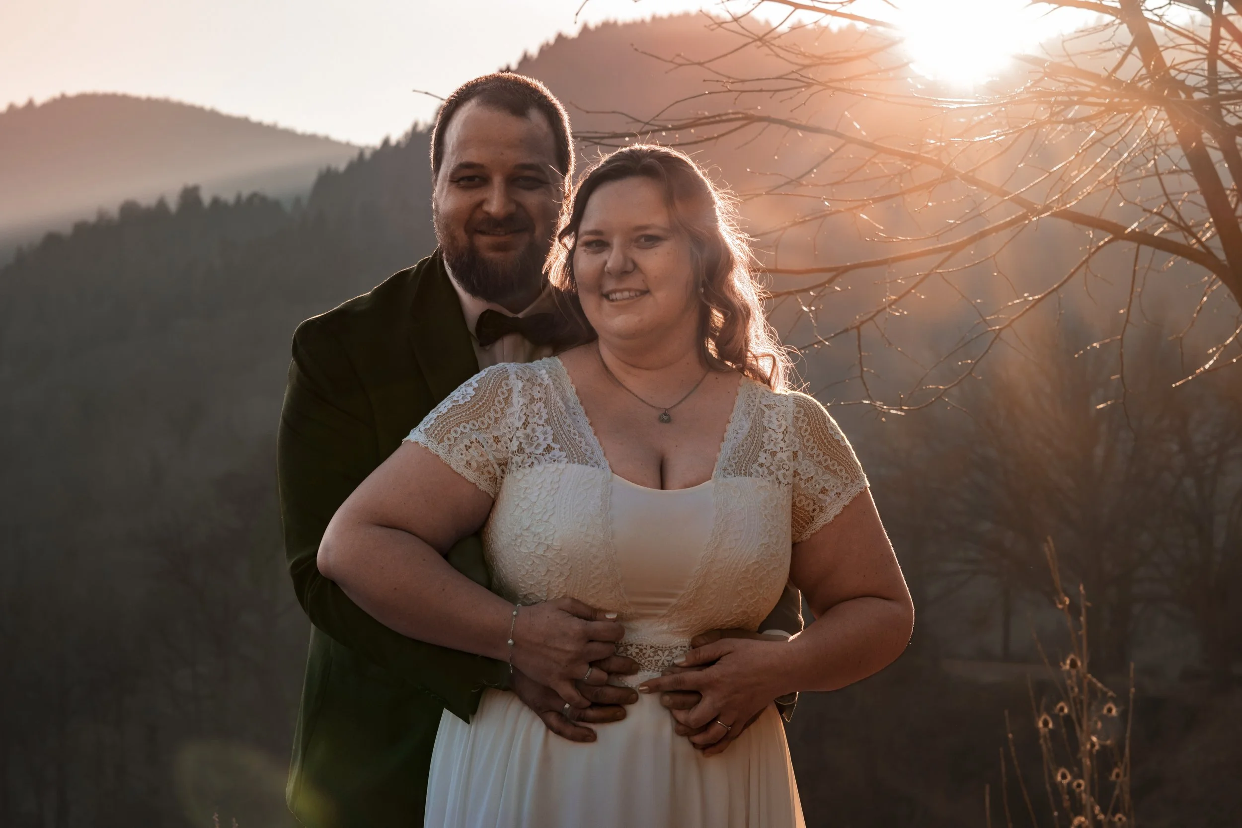 Ein Mann in Smoking und eine Frau in einem weißen Kleid posieren draußen bei Sonnenuntergang vor einer Berglandschaft.