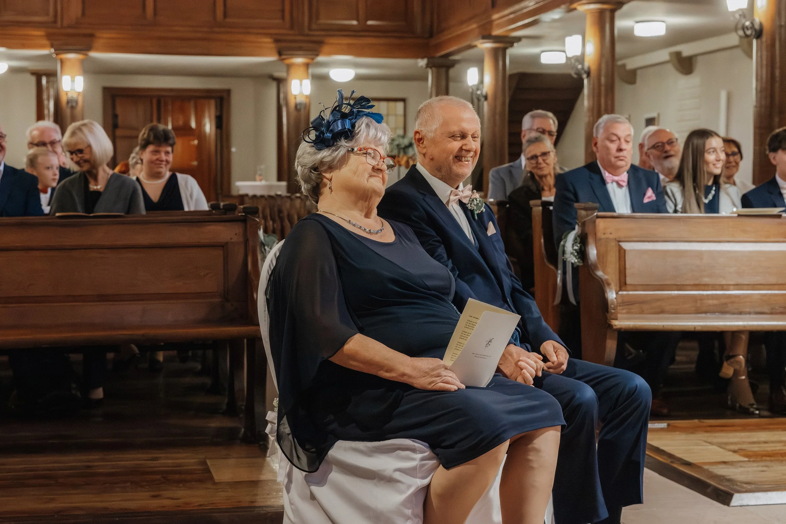 Eine Hochzeitsfeier in einer Kirche mit einem älteren Ehepaar im Vordergrund, das vermutlich die Eltern des Brautpaares sind. Beide tragen formelle Kleidung, die Frau trägt eine dunkle dress und eine blaue Hutdekoration, der Mann einen Anzug mit Flie