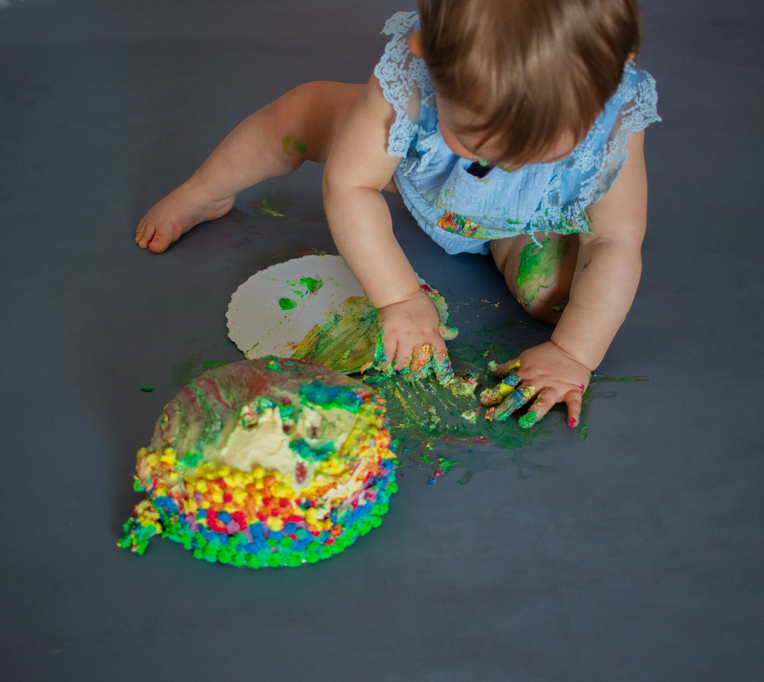 Ein kleines Kind, das auf einem dunklen Boden spielt, umgeben von buntem, zerbrochenem Geburtstagskuchen und buntem Zuckerguss. Das Kind trägt ein blaues Kleid mit Spitzen.