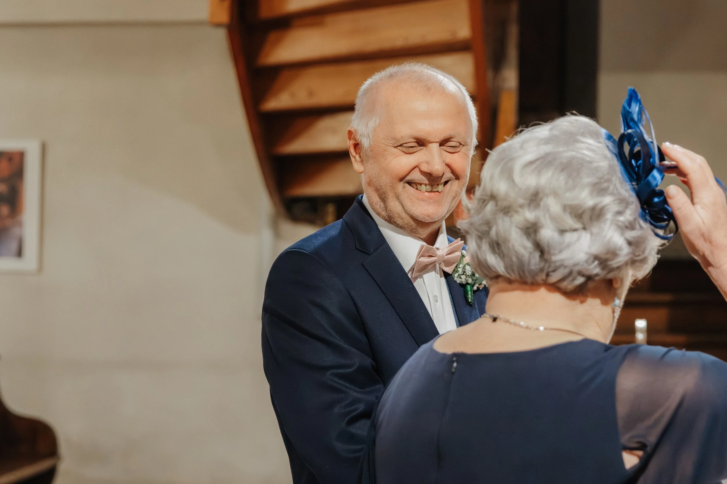 Ein lachender Mann in formeller Kleidung, vermutlich bei einer Hochzeit, mit einer älteren Frau, die ein Hut trägt, die anscheinend ihnen einen Blumenstrauß oder ähnliches anpasst.