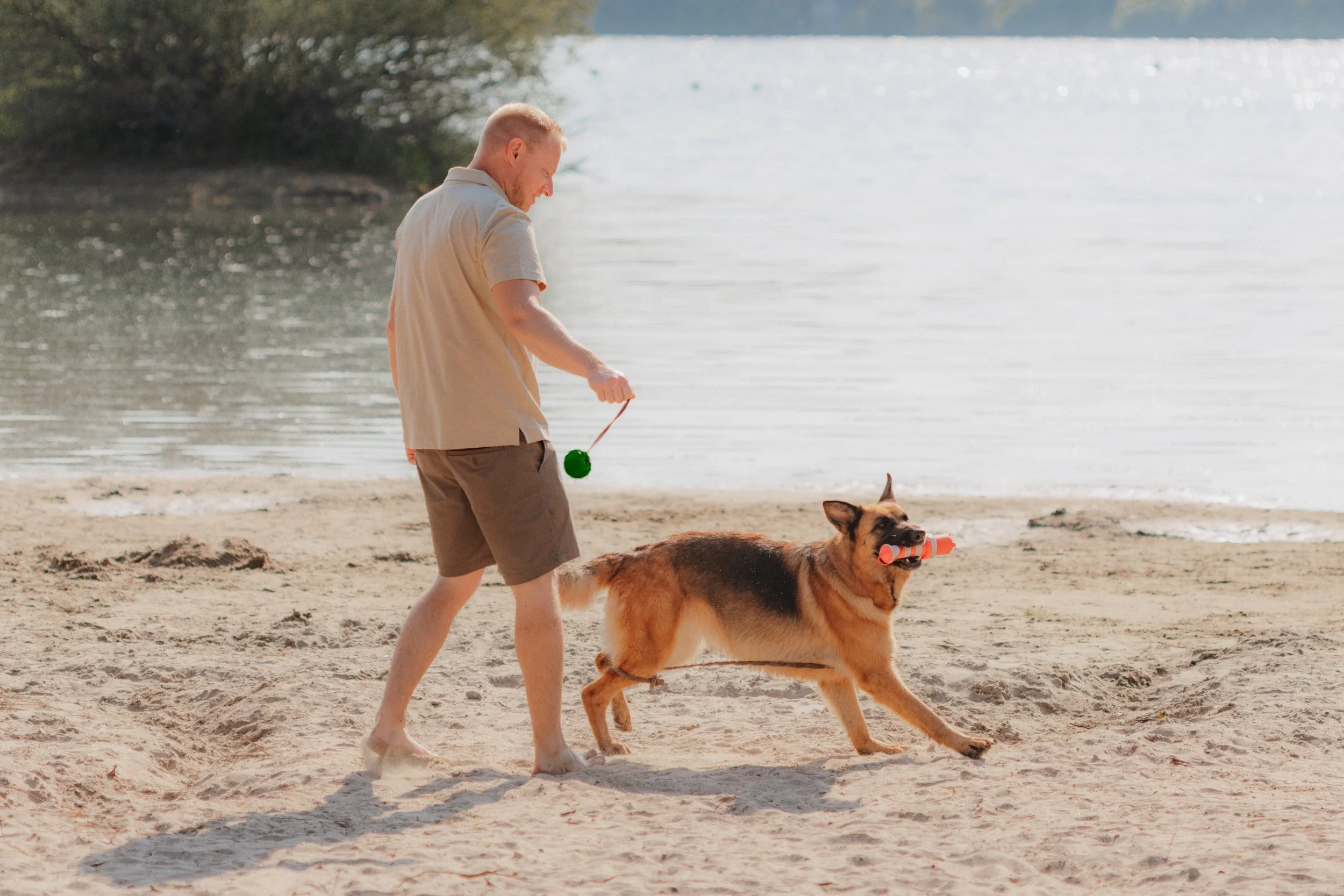 Ein Mann spielt mit einem Hund am Strand, beide spielen mit Spielzeug, der Hund trägt einen Gegenstand im Maul, im Hintergrund Wasser und Bäume.