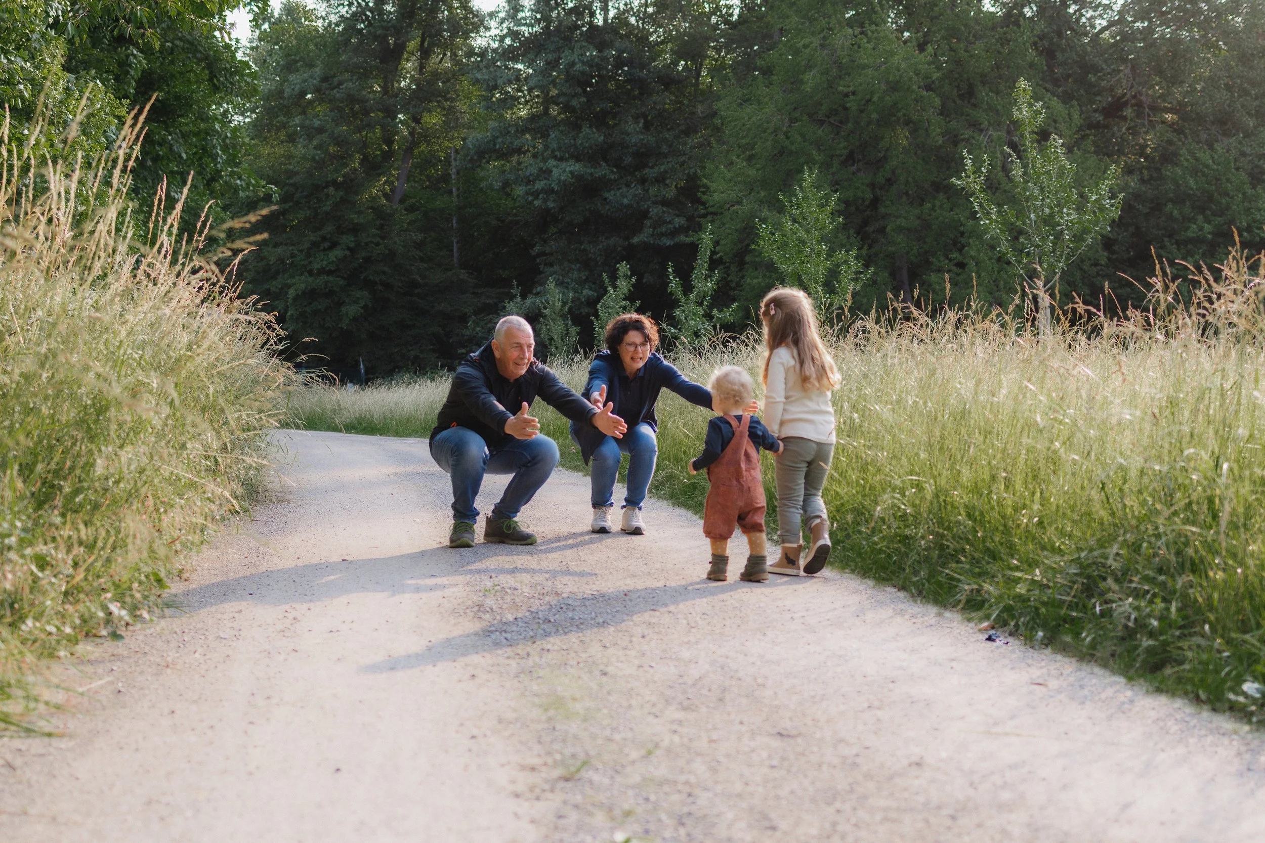 Eine Familie auf einer Parkstraße, bestehend aus zwei Erwachsenen und zwei Kindern, umarmt sich und genießt die Natur, umgeben von Büschen und Bäumen im Grünen.