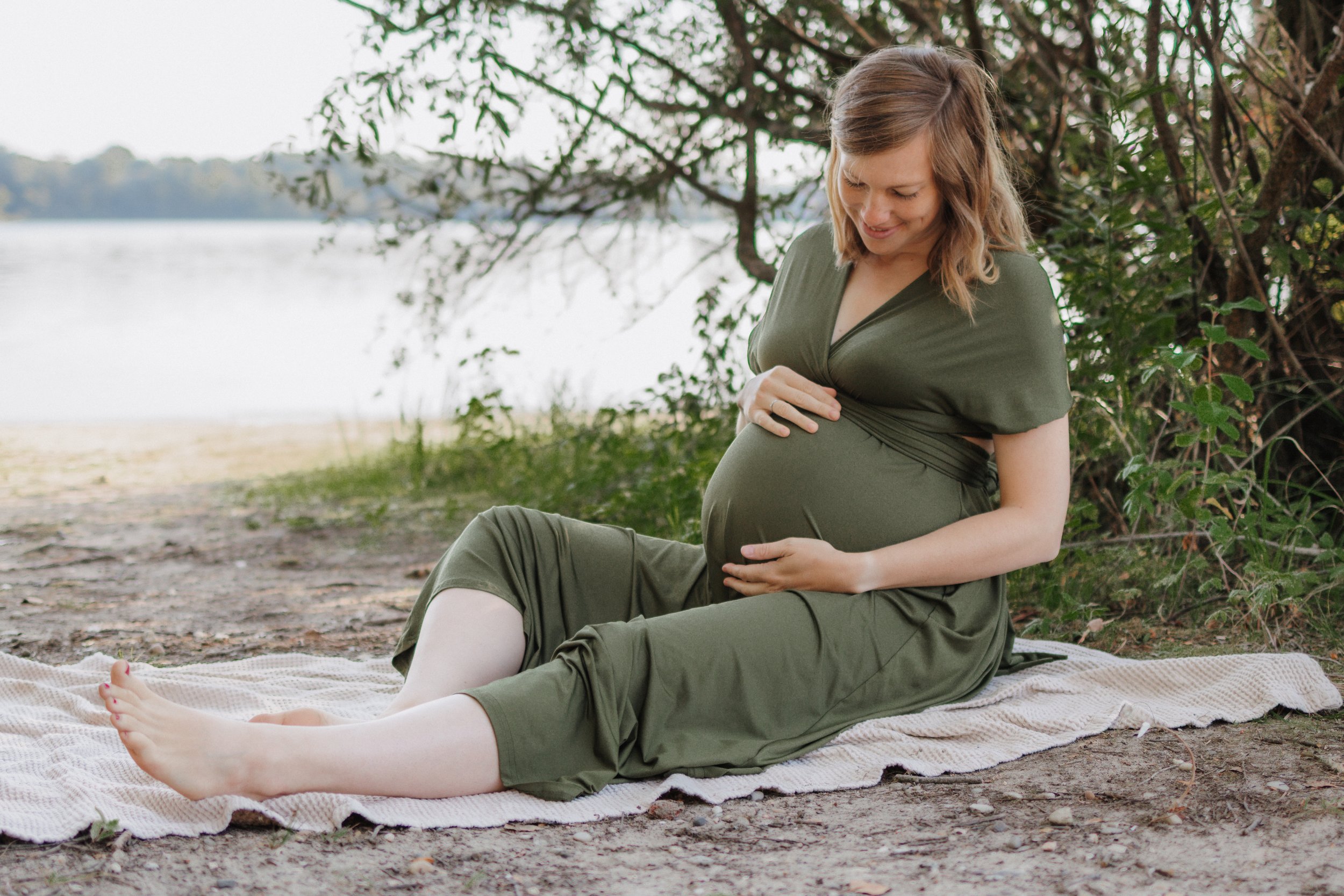 Schwangere Frau sitzt auf einer Decke am Flussufer im Grünen, lächelt und hält ihren Babybauch.