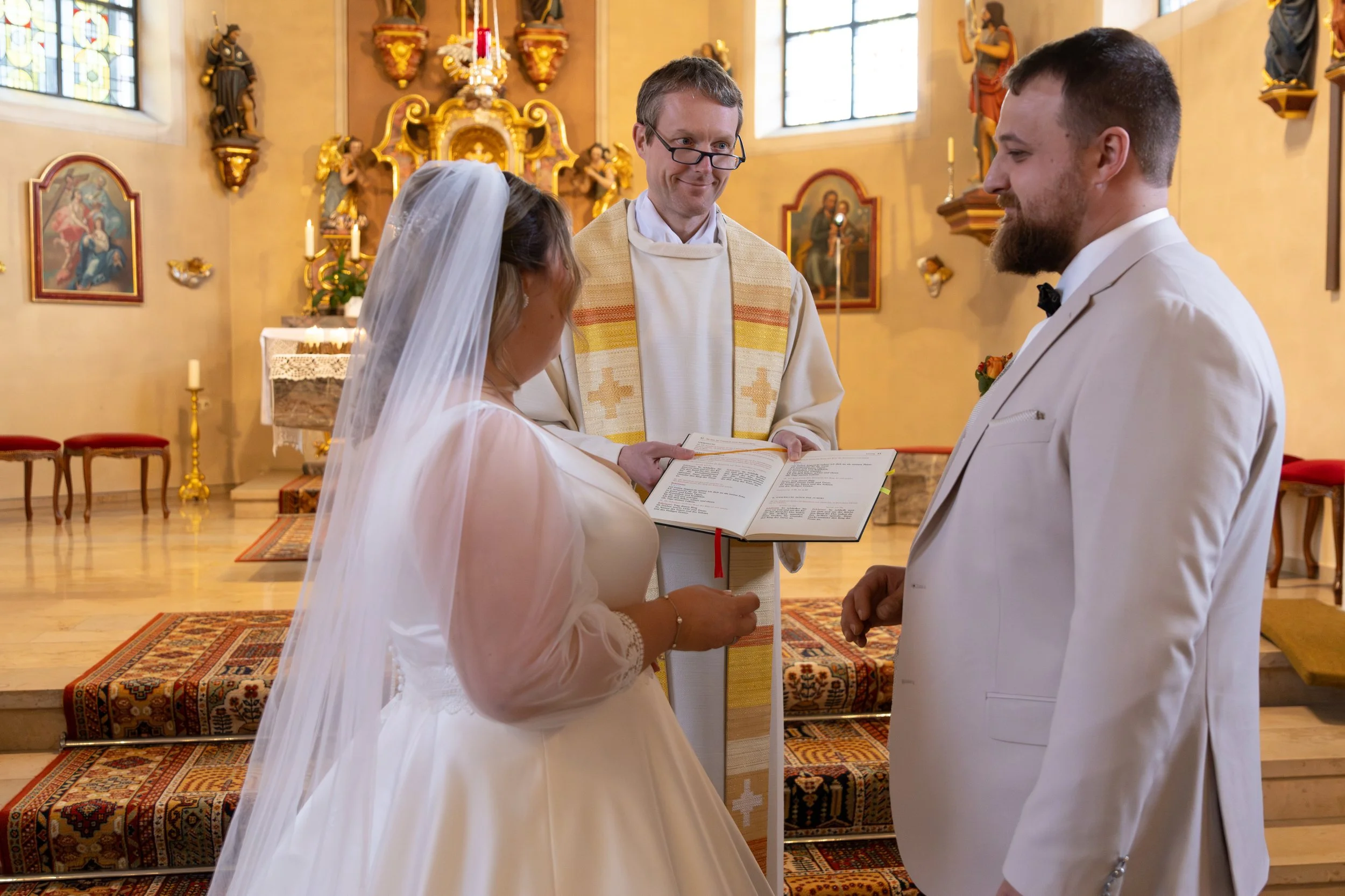 Ein Brautpaar steht bei einer Heirat in einer Kirche, die Braut trägt ein weißes Kleid und einen Schleier, der Bräutigam trägt einen weißen Anzug, der Priester hält ein Buch und steht vor ihnen.