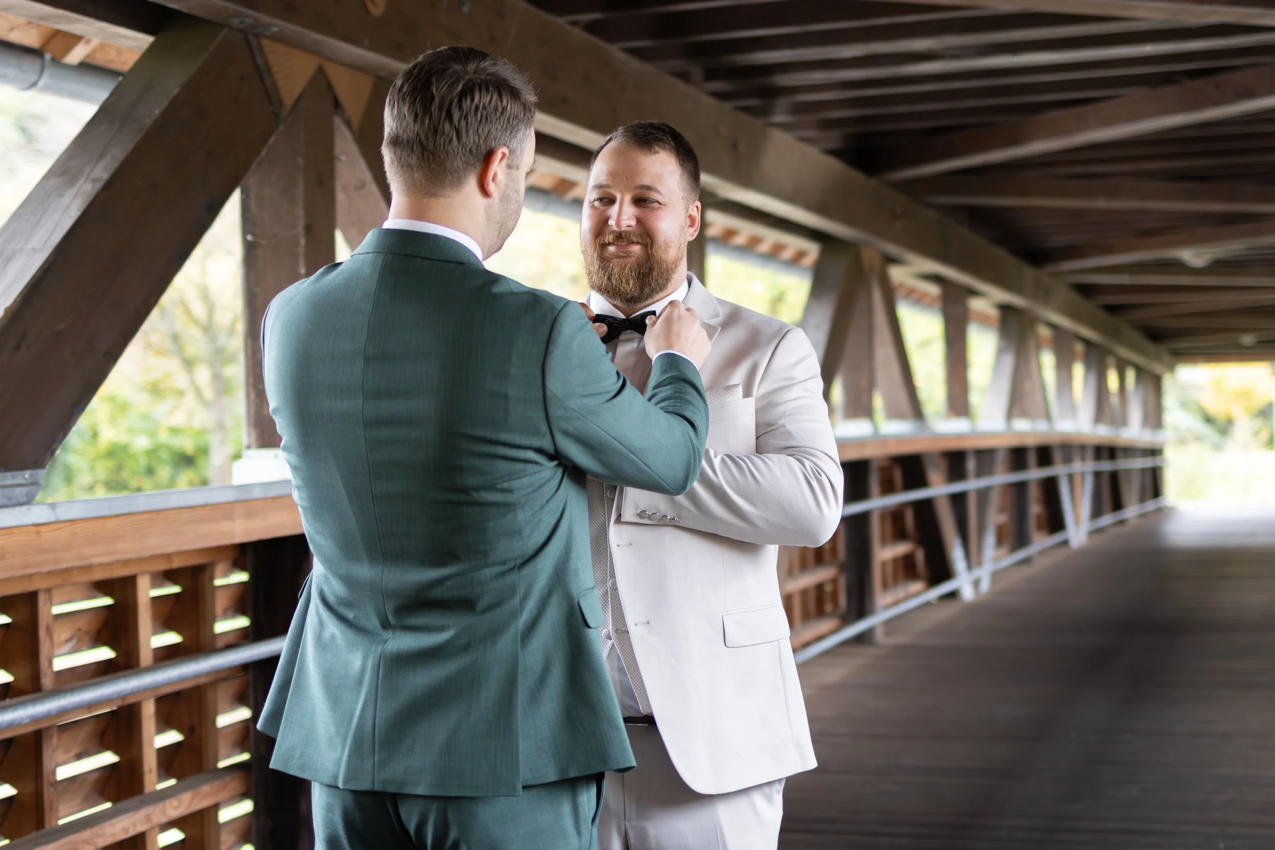 Zwei Männer in Anzügen auf einer Brücke, einer hilft dem anderen, den Krawattenknoten zu binden.