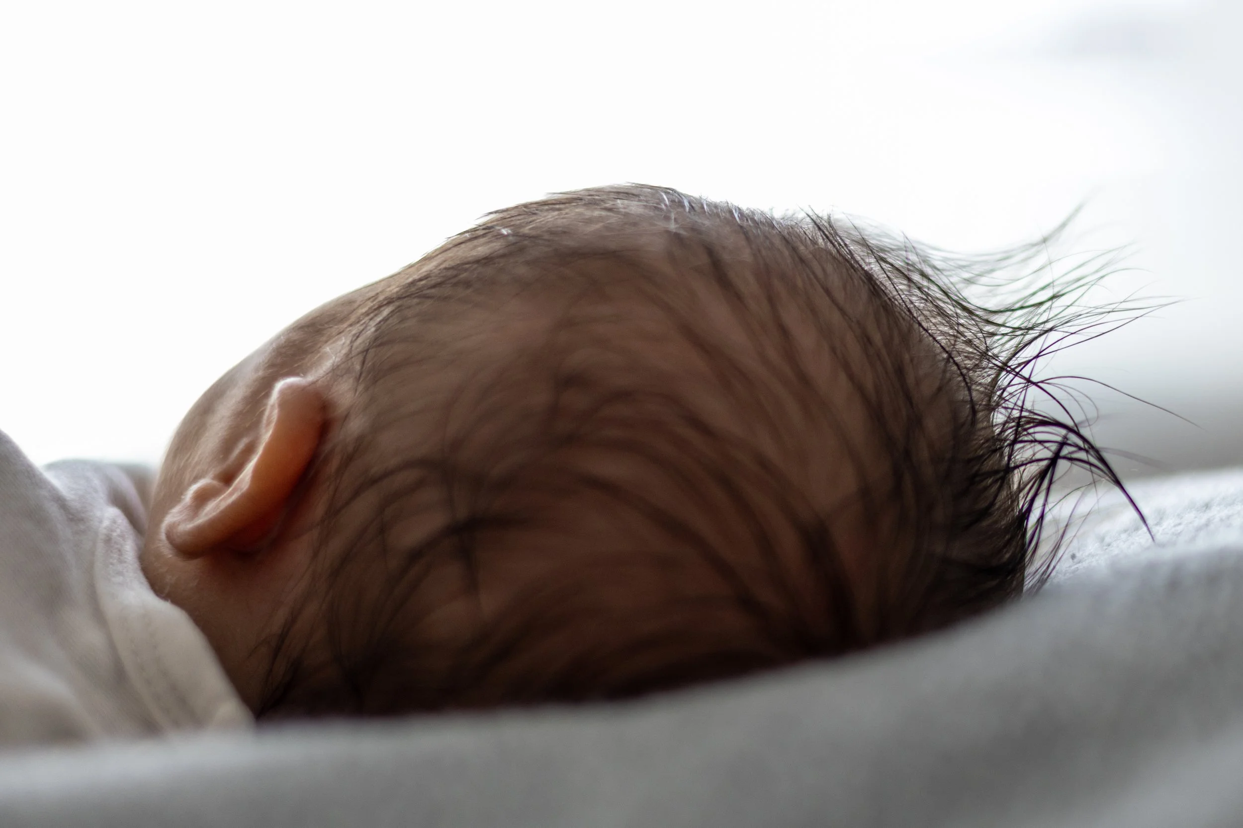 Nahaufnahme eines schlafenden Babys mit nacktem Kopf, liegend auf einer Oberfläche, mit Fokus auf Kopf und Haar, im weichen Licht.