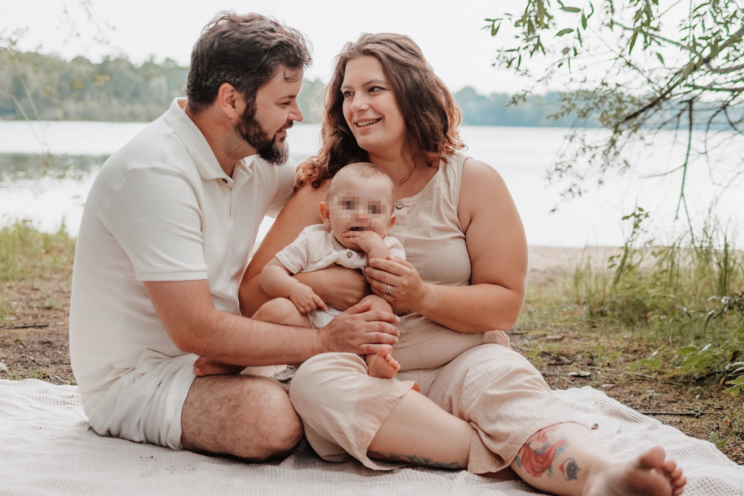 Familie sitzt am Ufer eines Sees im Freien, lachend und spielend, Kind mit Pixelausschnitt auf dem Gesicht, im Hintergrund Bäume und Wasser.