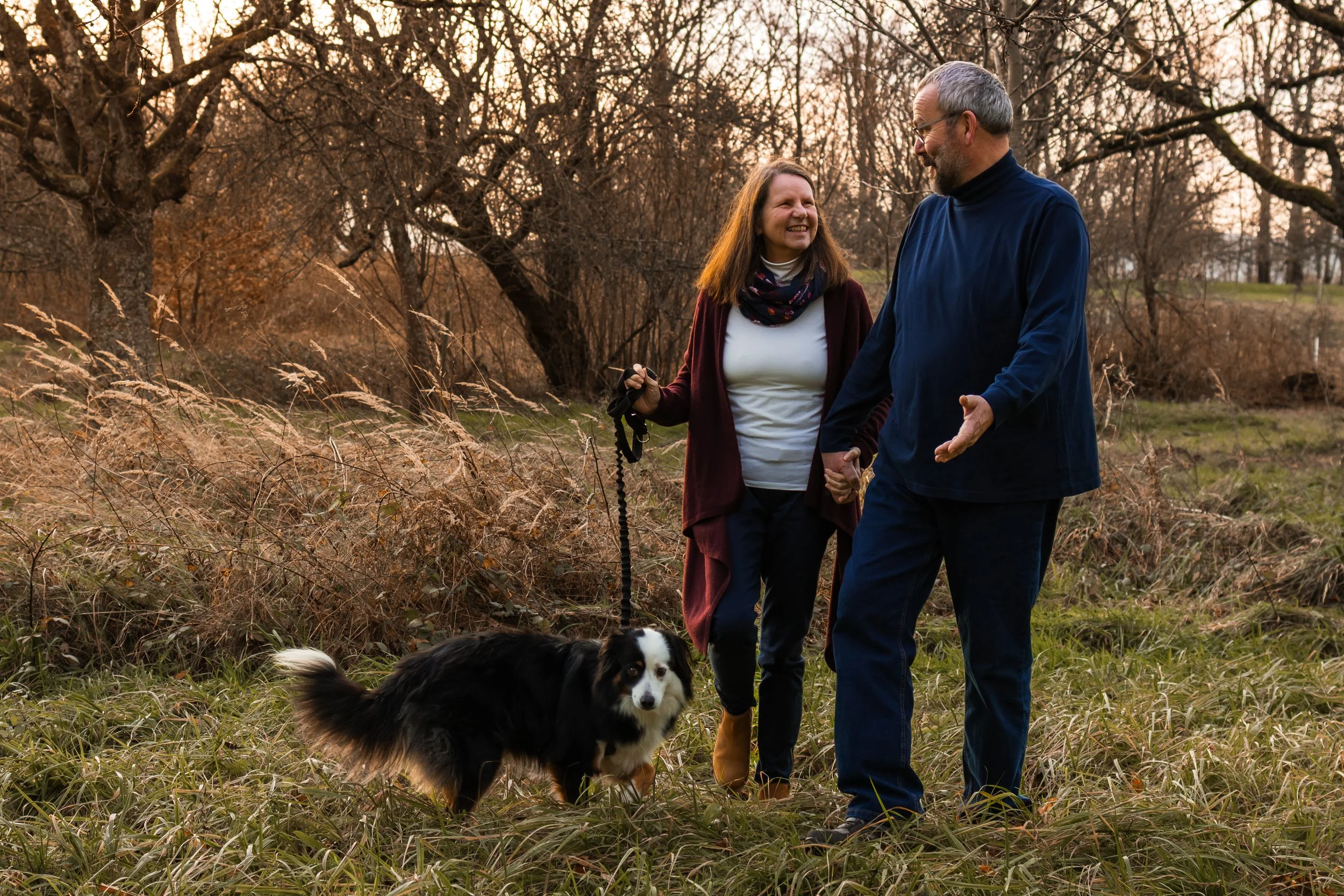 Ein Mann und eine Frau, die im Sonnenuntergang im Freien spazieren gehen, Händchen haltend, mit einem Hund an der Leine, umgeben von Bäumen und Gras
