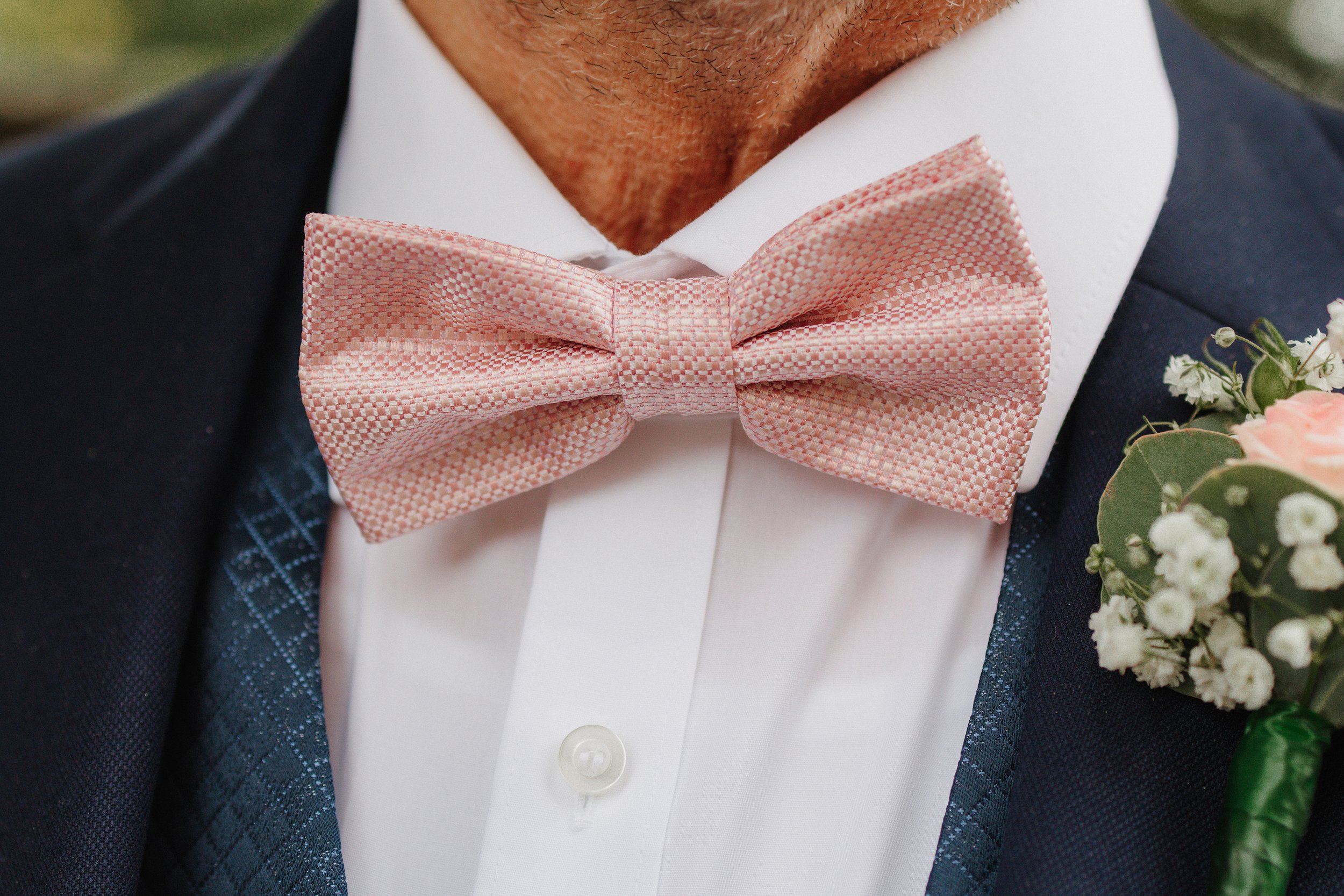 Nahaufnahme eines Mannes, der einen weißen Hemd, einen schwarzen Anzug und eine rosa Fliege trägt, mit einem kleinen Blumenstrauß an der Brust.