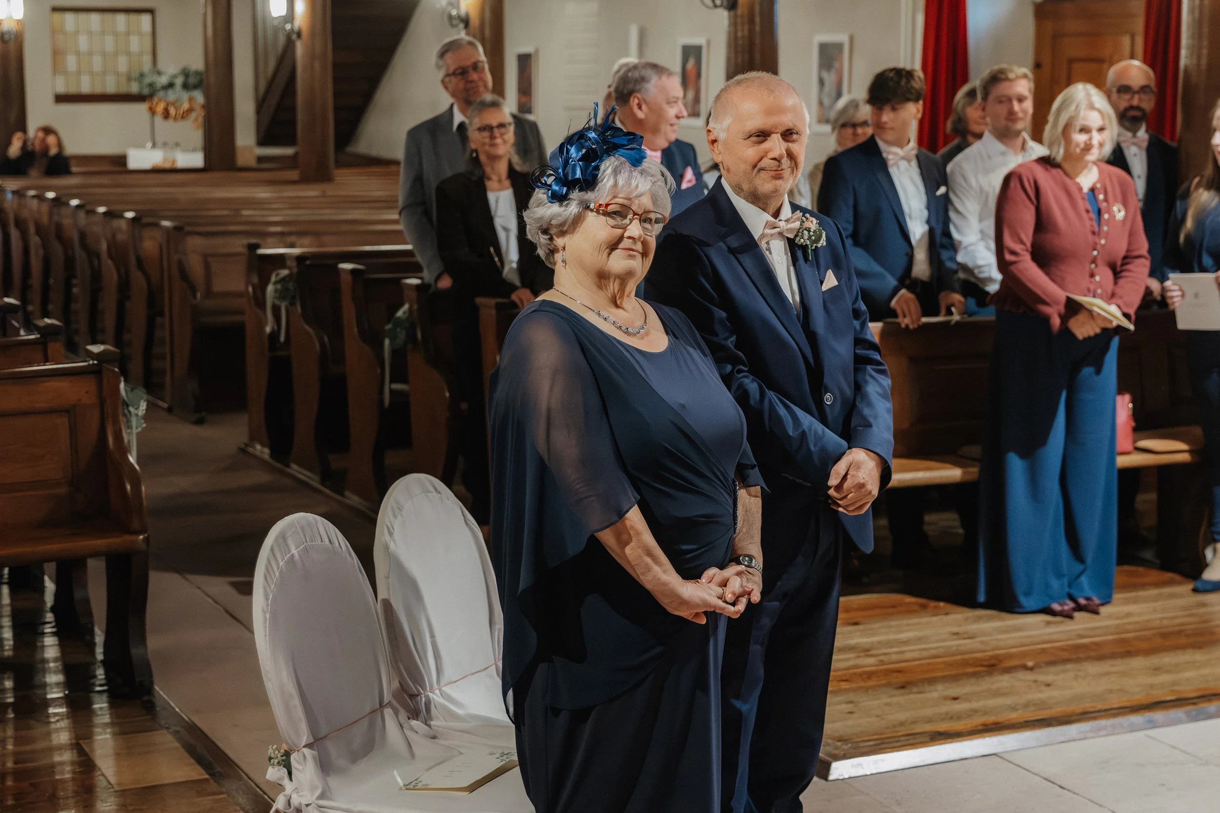 Ein älteres Paar bei einer Hochzeit in einer Kirche, beide tragen formelle Kleidung, die Frau trägt eine dunkle Abendrobe mit Schmuck und einem Hut mit Schleife, der Mann trägt einen dunklen Anzug mit Fliege, im Hintergrund sitzen und stehen weitere 