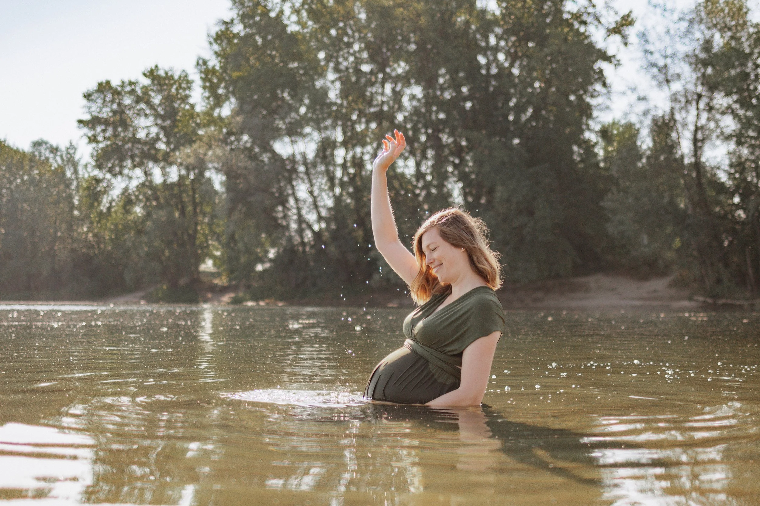 Schwangere Frau steht im Fluss, hebt die Hand und lächelt bei Sonnenuntergang, umgeben von Bäumen.