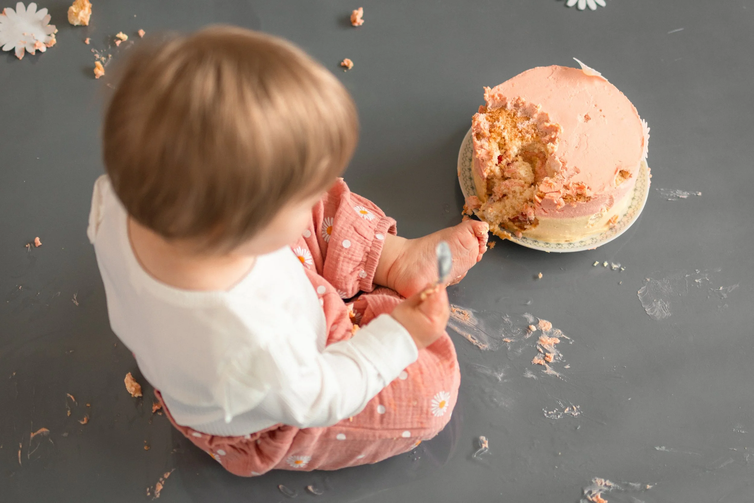 Kleines Kind sitzt auf dem Boden und isst Kuchen, während Krümel und ein Messer um ihn herum verstreut sind.
