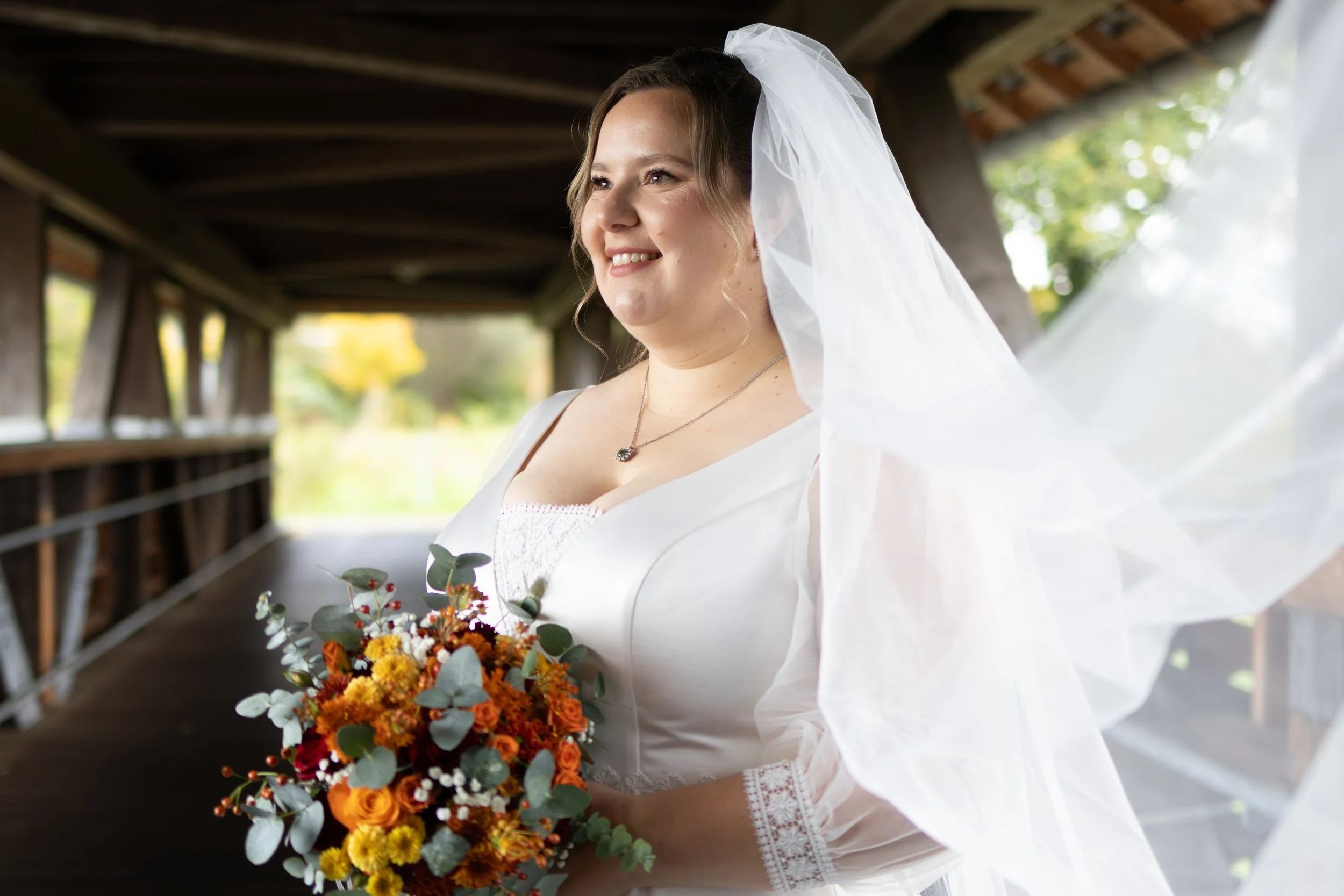 Braut in weißem Kleid mit Schleier, hält einen bunten Blumenstrauß, im Hintergrund eine Holzbrücke und Herbstbäume.