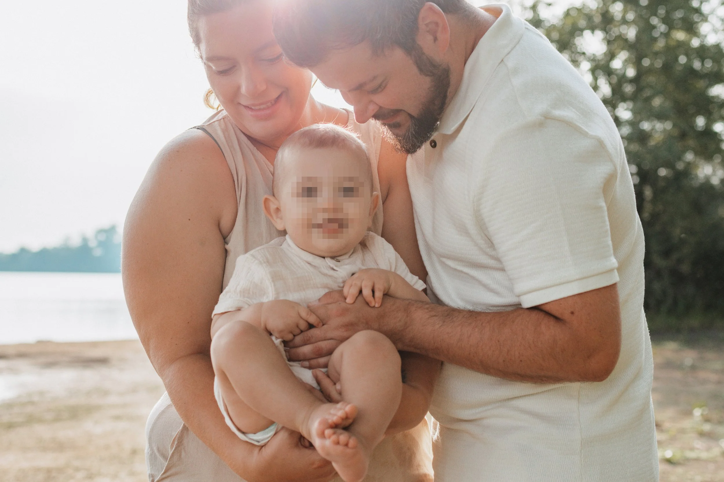 Ein glückliches Familienfoto von einem Paar, das einen kleinen Jungen am Strand hält, bei Sonnenuntergang, mit Bäumen im Hintergrund.