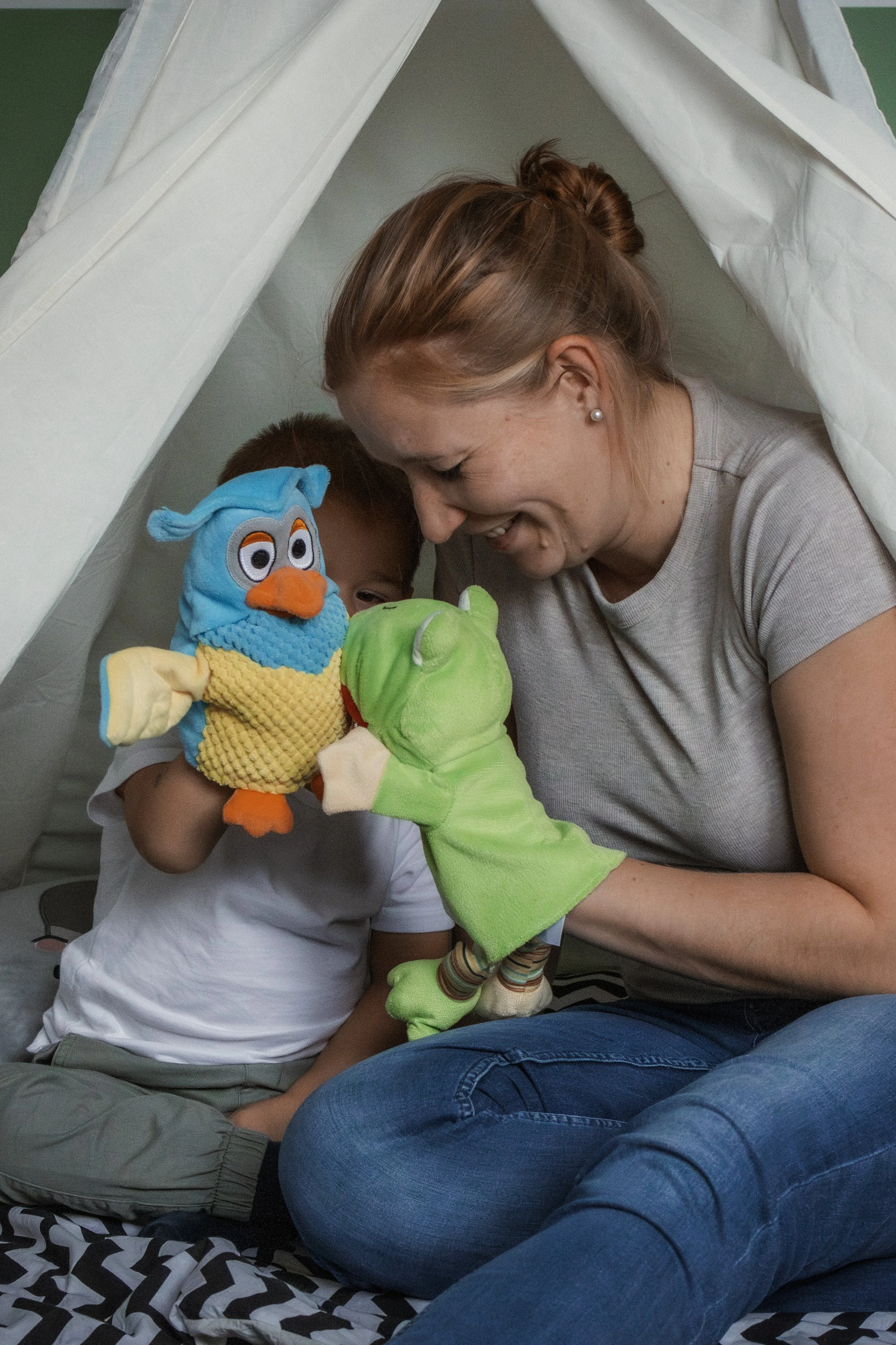 Eine Frau und ein Kind spielen mit Handpuppen unter einem Zelt. Die Frau hat braune Haare, trägt ein graues T-Shirt und blaue Jeans. Das Kind trägt ein weißes T-Shirt und graue Sweatpants. Sie verwenden eine blaue Vogel-Handpuppe und eine grüne Kroko