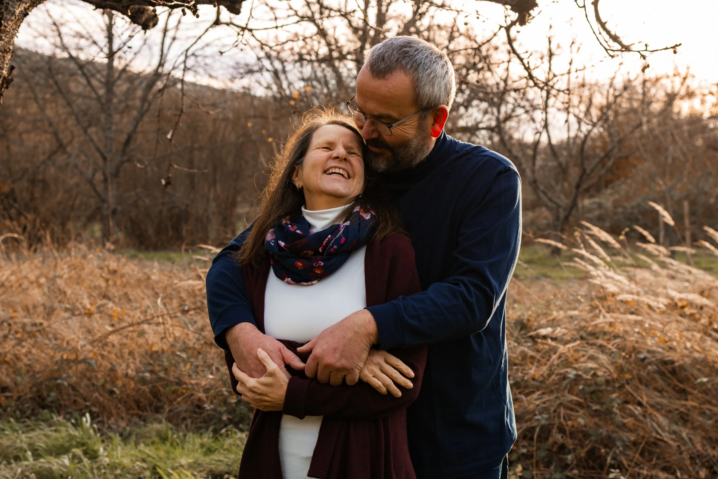 Ein älteres Paar umarmt sich lachend im Freien bei Sonnenuntergang, umgeben von Herbstlaub.