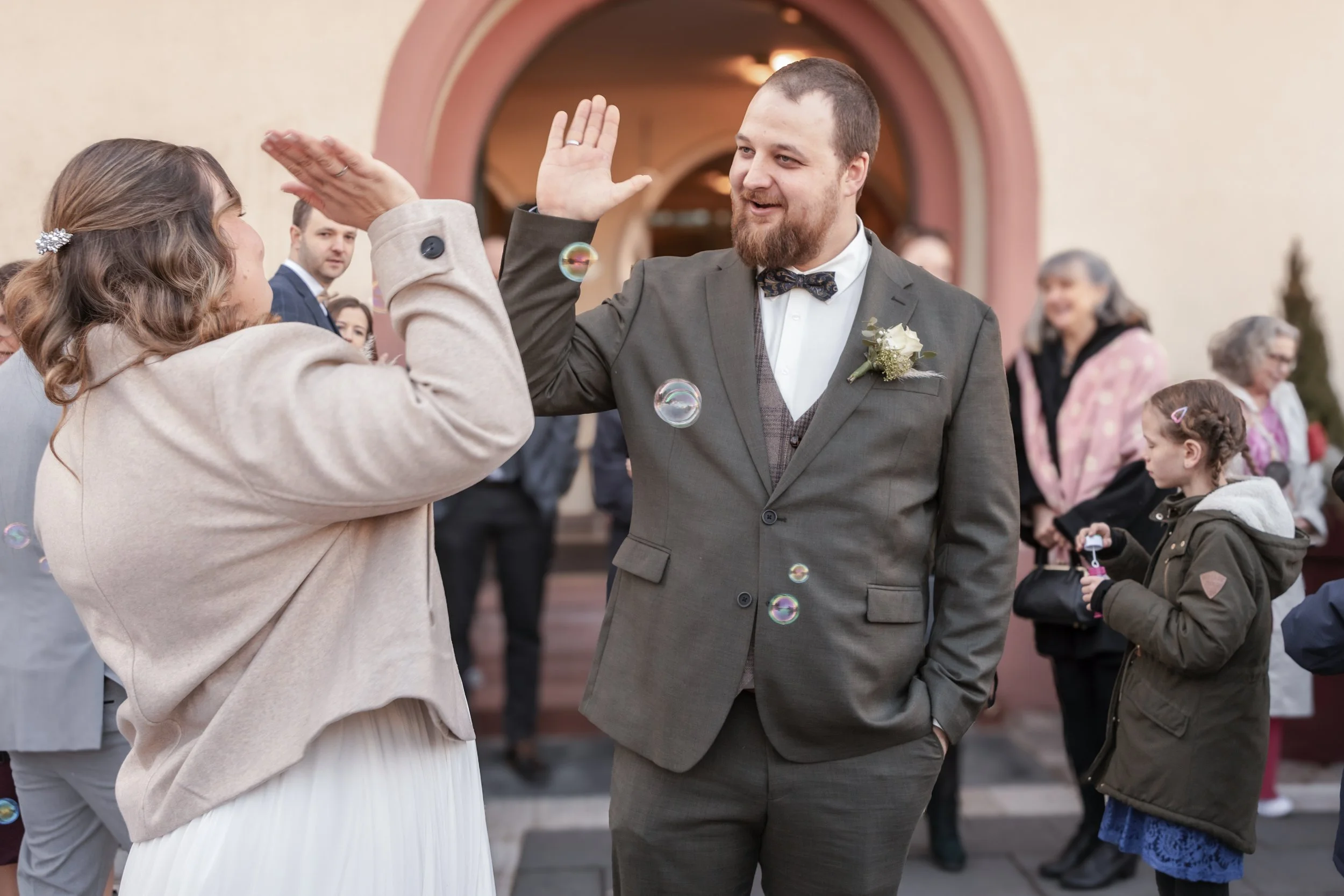 Ein Mann und eine Frau beim High-Five vor einer Kirche, umgeben von Gästen bei einer Hochzeit.