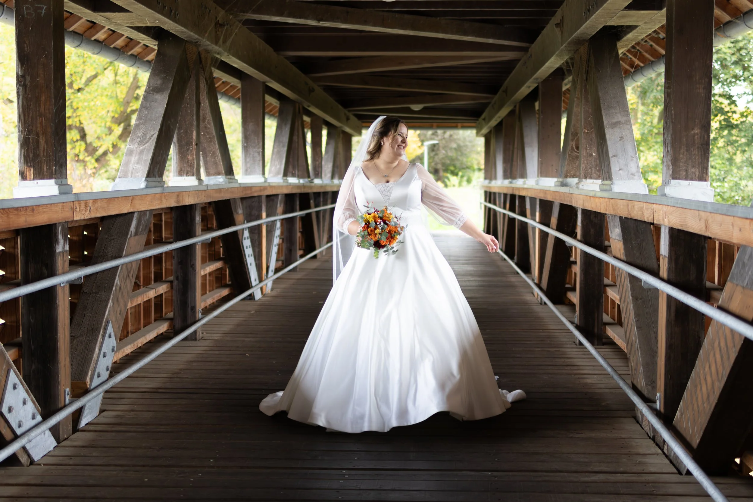 Braut in weißem Hochzeitskleid mit Blumenstrauß auf einer Holzzugbrücke unter einem Holzdach im Freien.